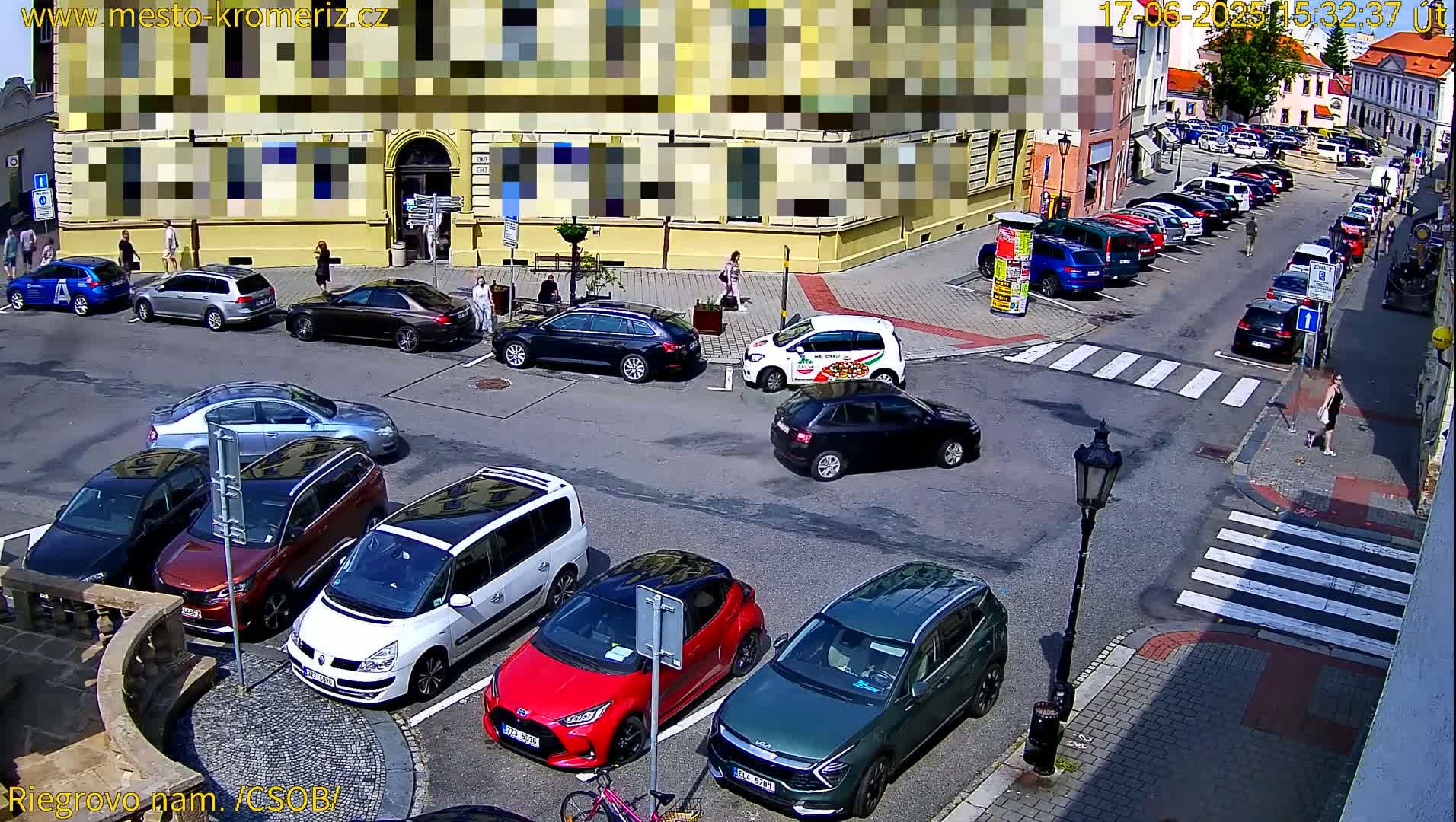 A sunny city square with parked cars and pedestrians, some cars are driving through the intersection.
