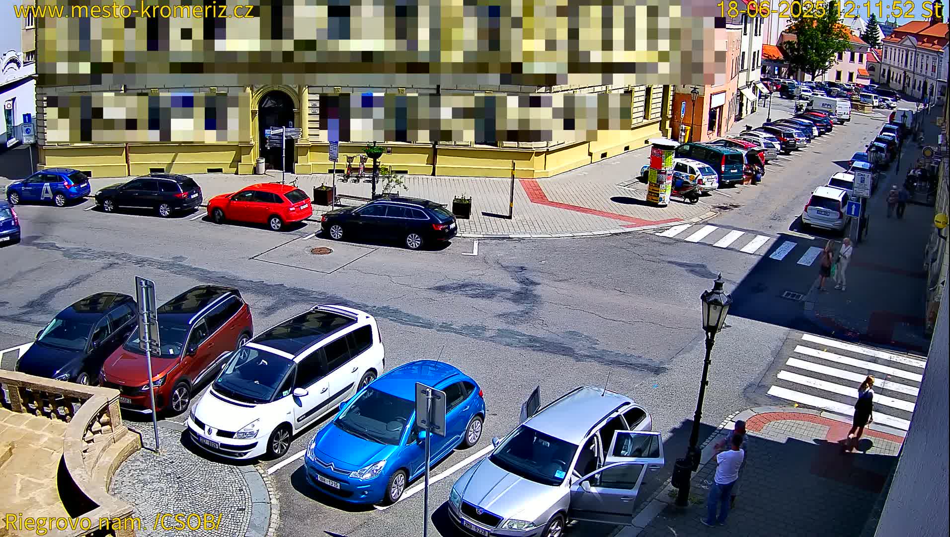 A sunny street corner with several cars parked and a few people walking on the sidewalk.