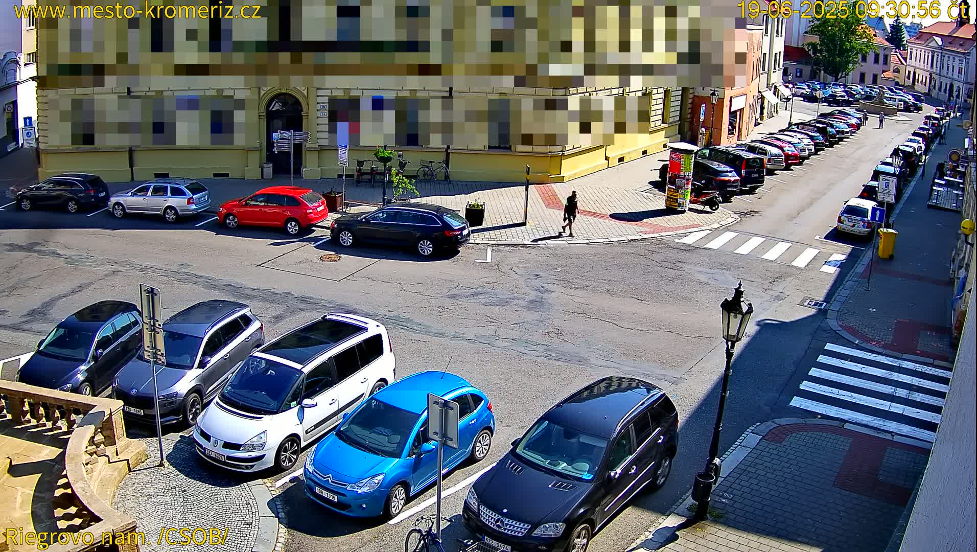 A sunny street corner shows several parked cars, a person walking, and a building with its windows obscured.