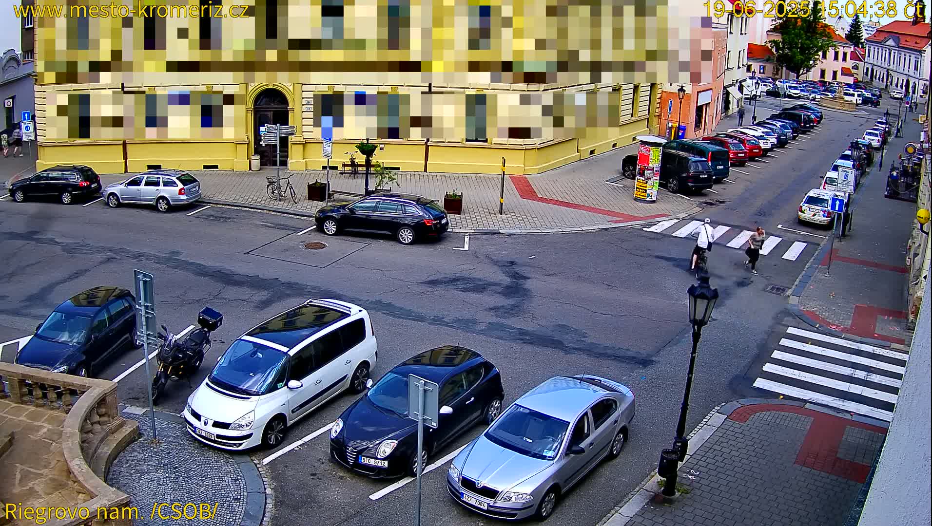 A street corner view showing several parked cars and a few pedestrians crossing a crosswalk, on a sunny day.
