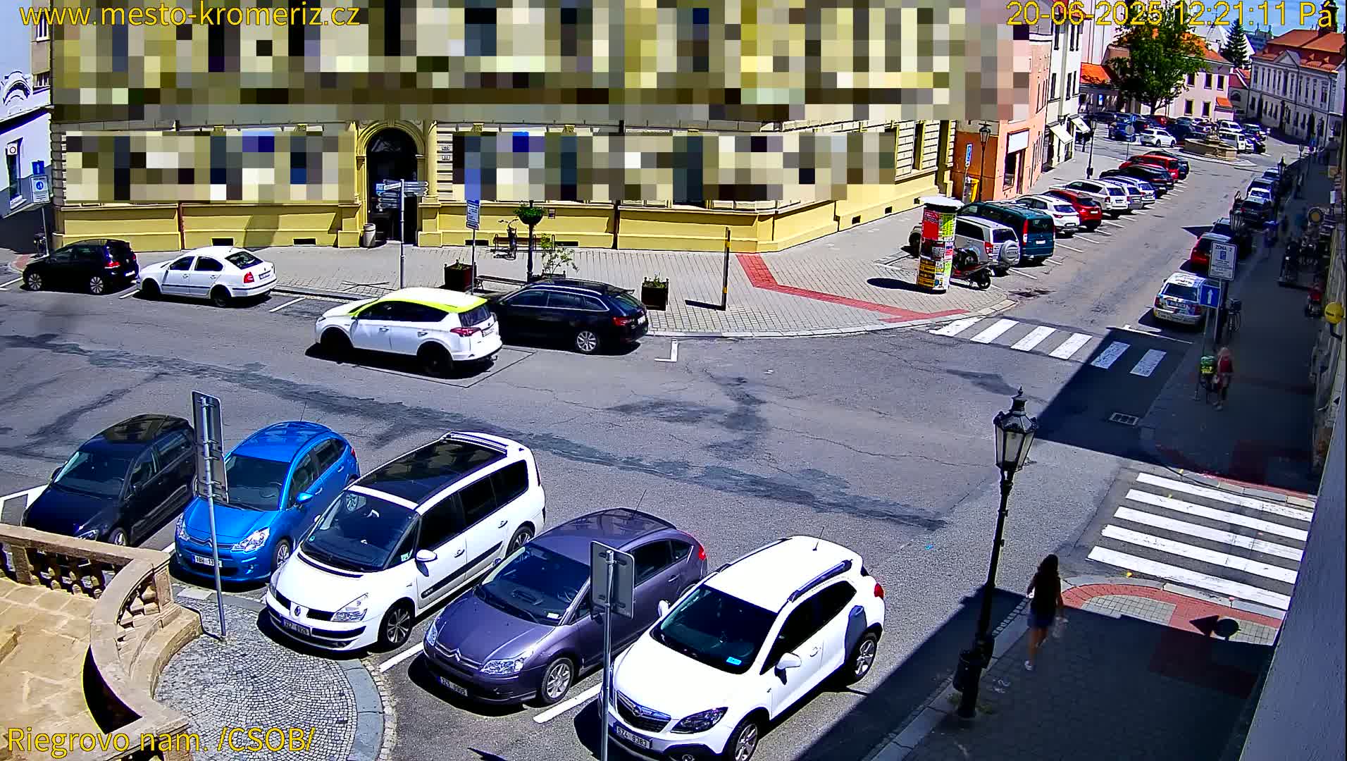 A sunny day shows several cars parked in a lot and along a street near a building, with a pedestrian crossing and a few pedestrians visible.