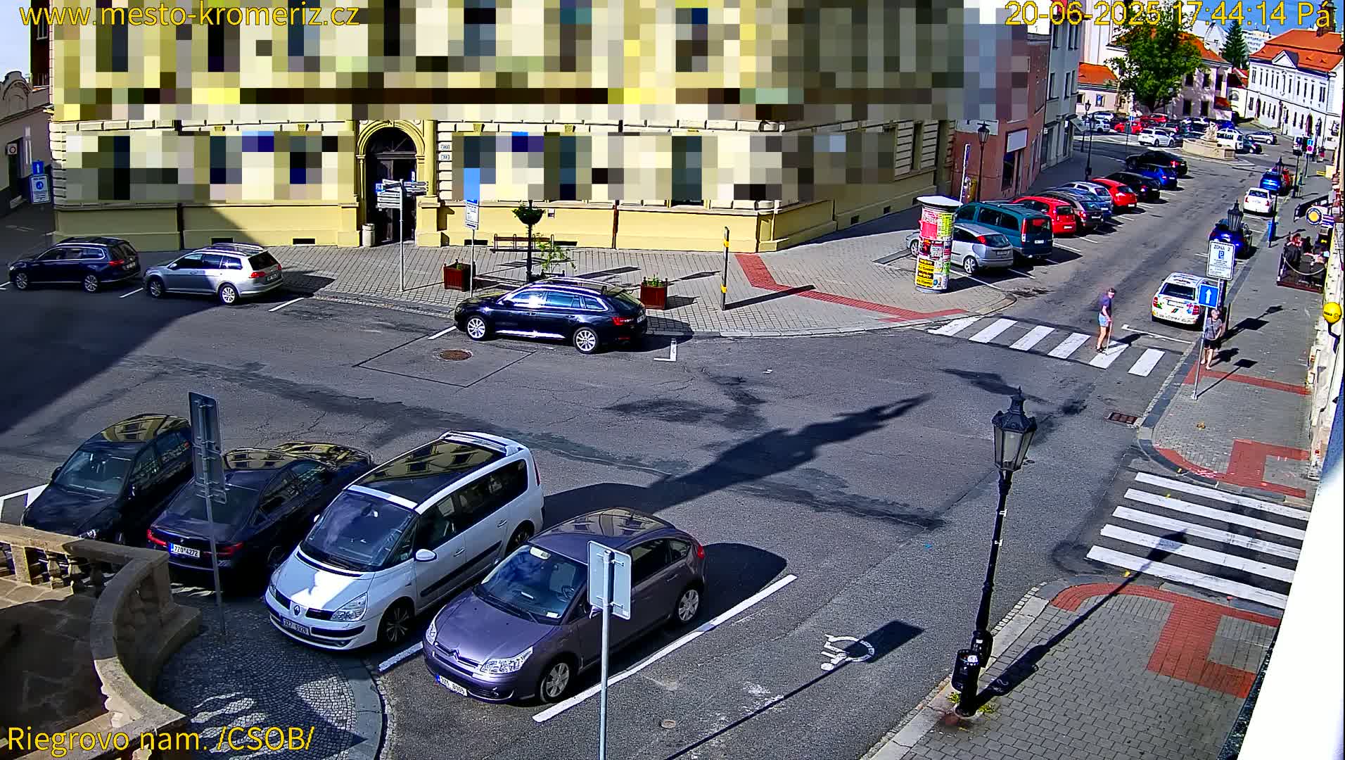Several cars are parked in a sunny city parking lot near a crosswalk where two pedestrians are walking.