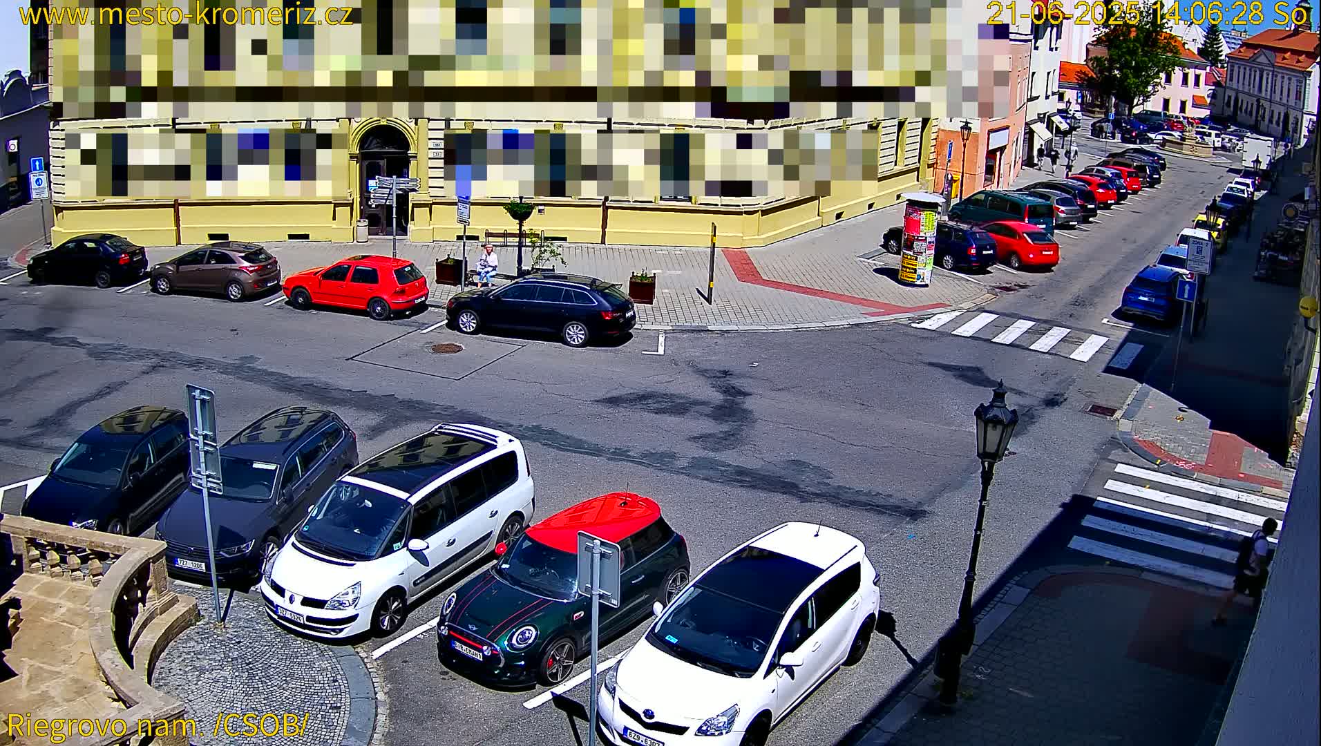 Several cars are parked on a sunny street in front of a yellow building, with more cars parked further down the street.
