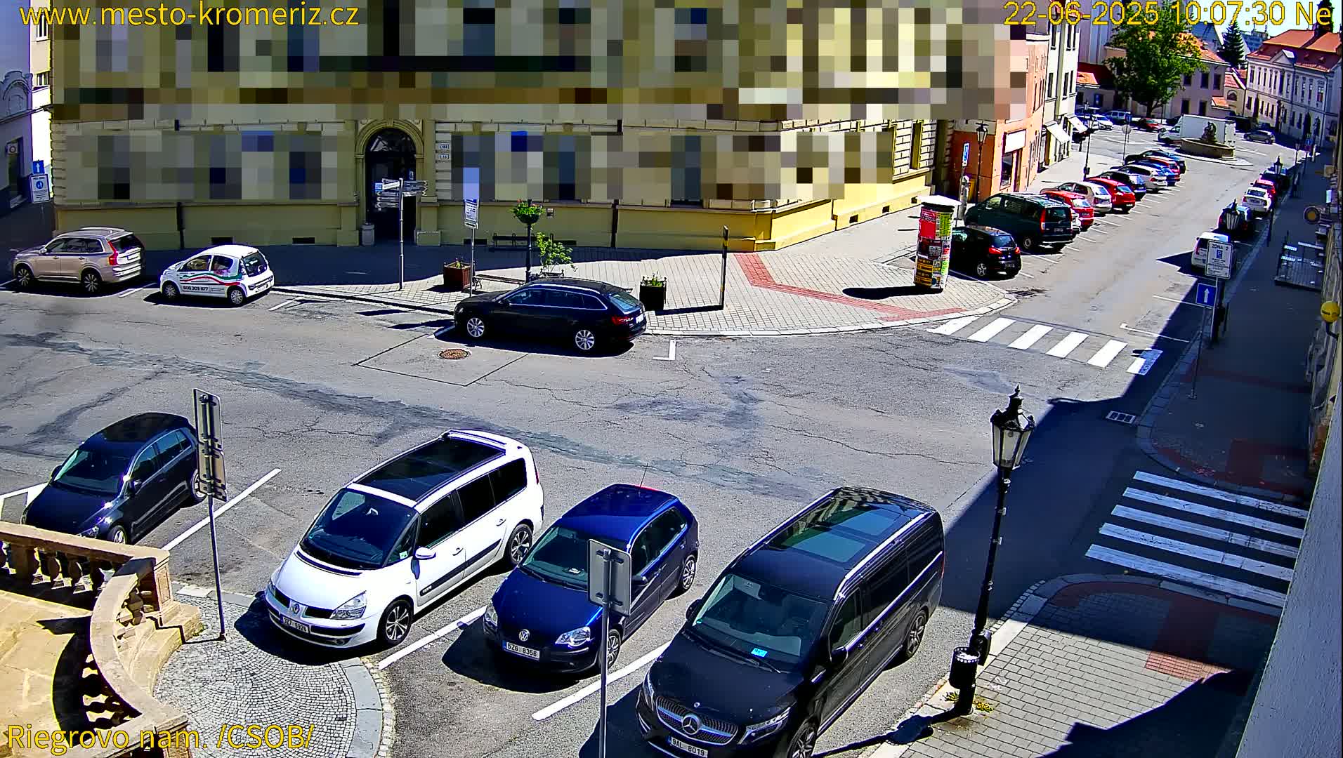 A sunny street scene shows several cars parked in a lot and along a curb next to a yellow building.