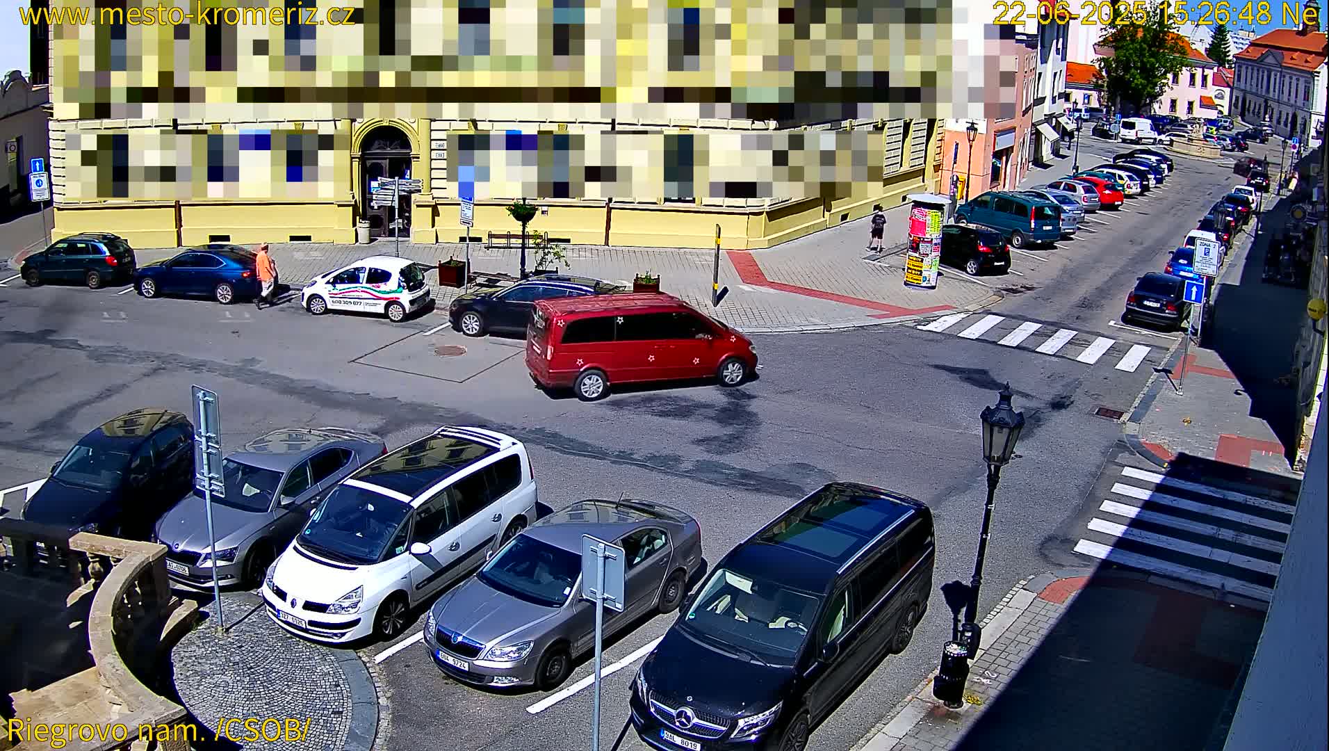 A sunny street scene shows several cars parked along the curb and a red minivan in the middle of a crosswalk, with a yellow building in the background.