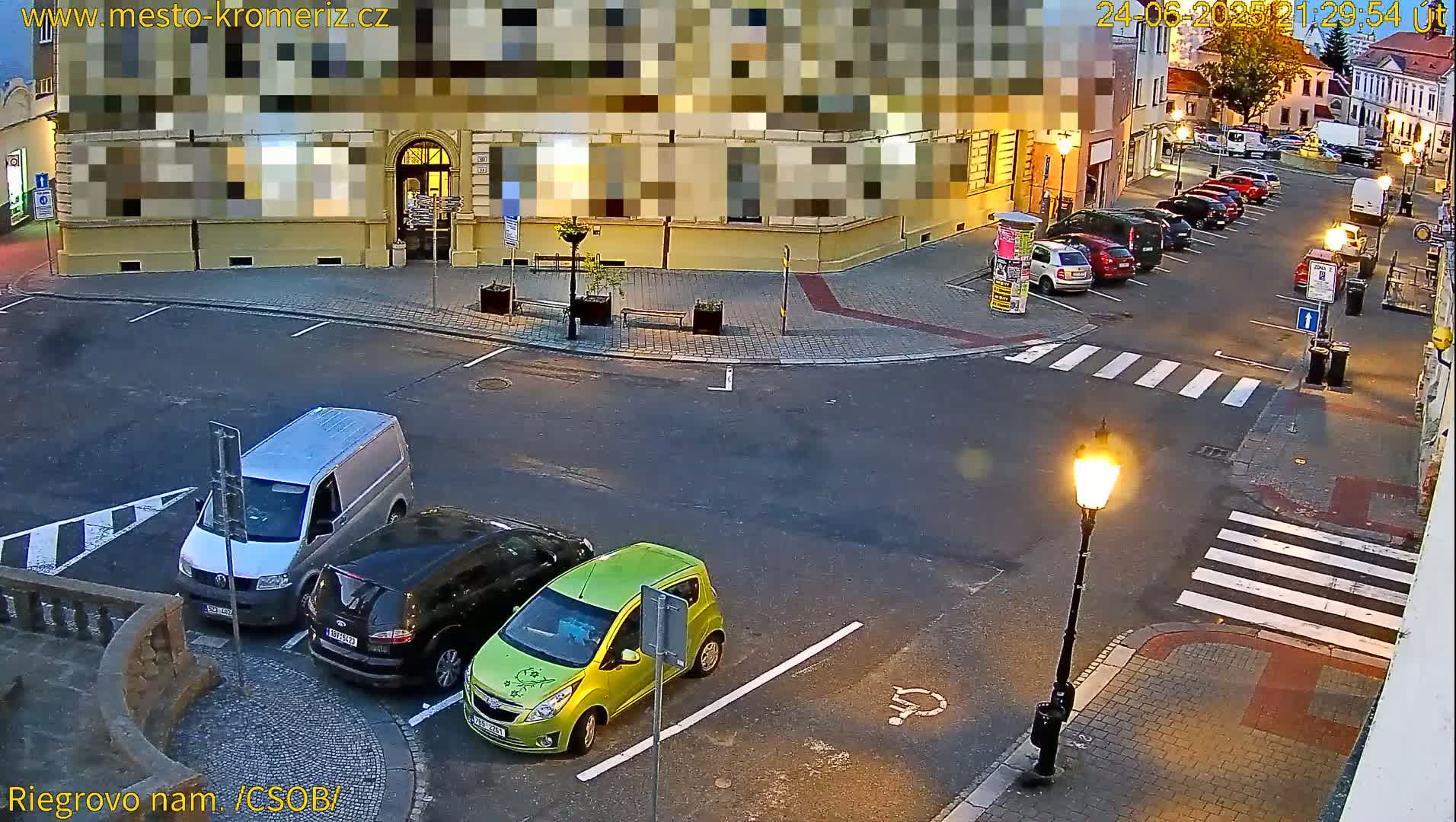 A street corner at dusk with several parked cars, a van, and a streetlamp, under partly cloudy skies.