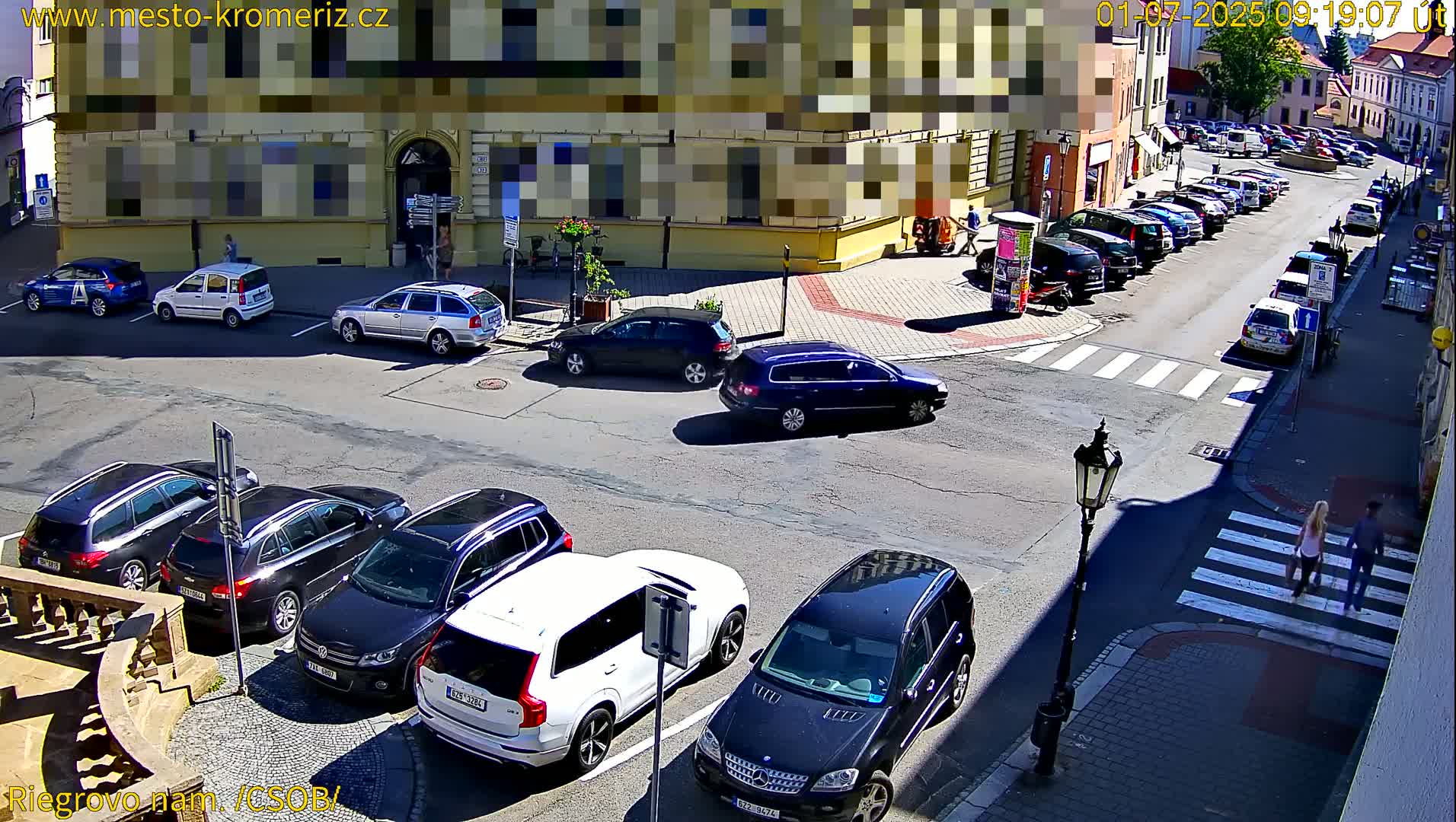 A sunny street corner shows several parked cars and a few cars in motion, with a crosswalk and pedestrians visible.