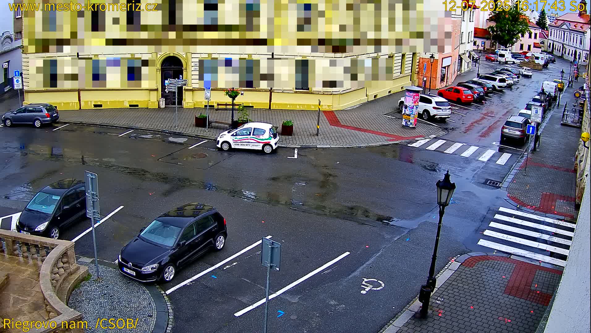 A sunny street corner shows several parked cars and a few cars in motion, with a crosswalk and pedestrians visible.