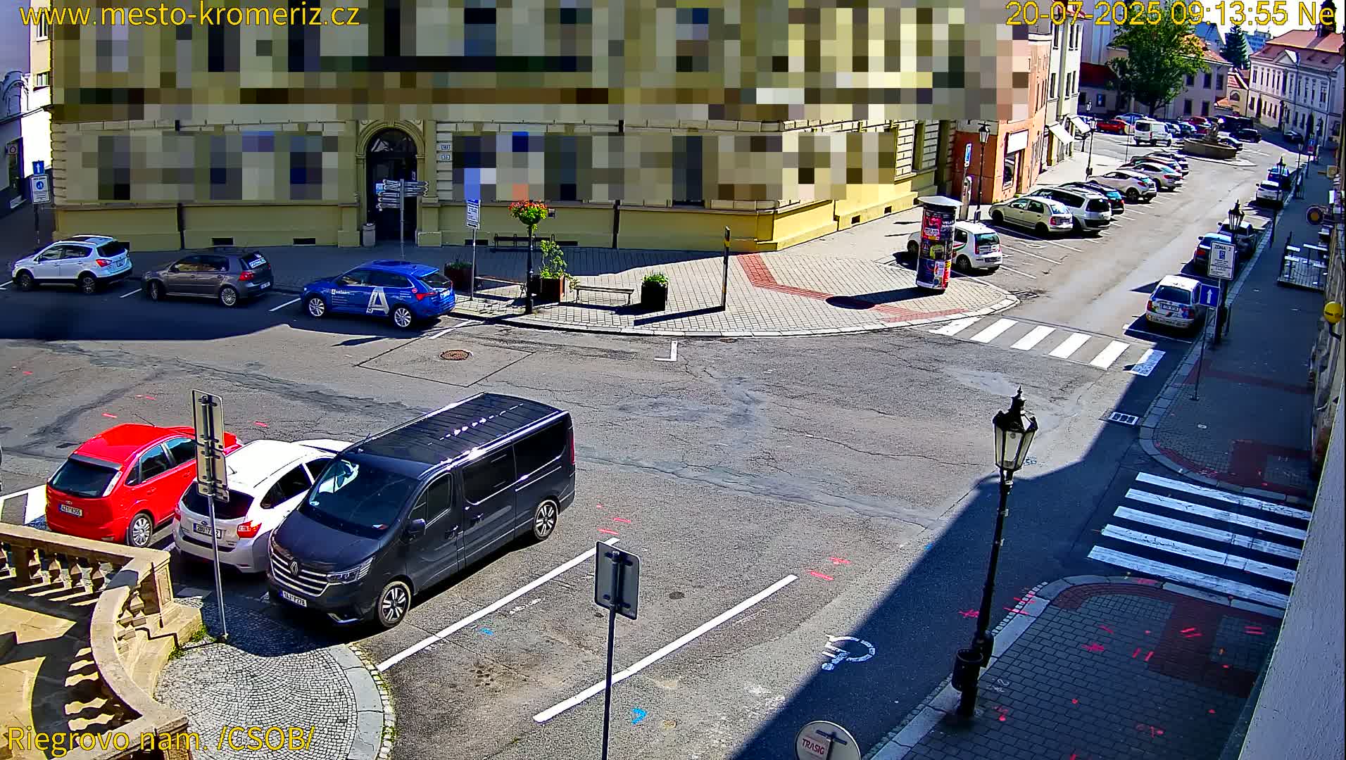 A sunny street corner shows several parked cars and a minivan, with more cars parked along a side street.