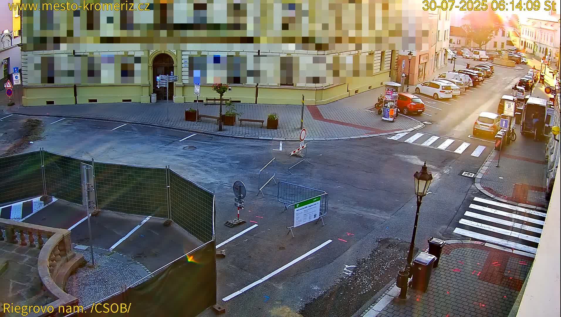 A sunny street corner shows several parked cars and a minivan, with more cars parked along a side street.
