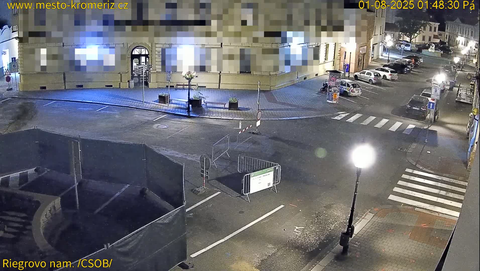 A nighttime view of a mostly empty town square with several parked cars, construction barriers, and a streetlamp.