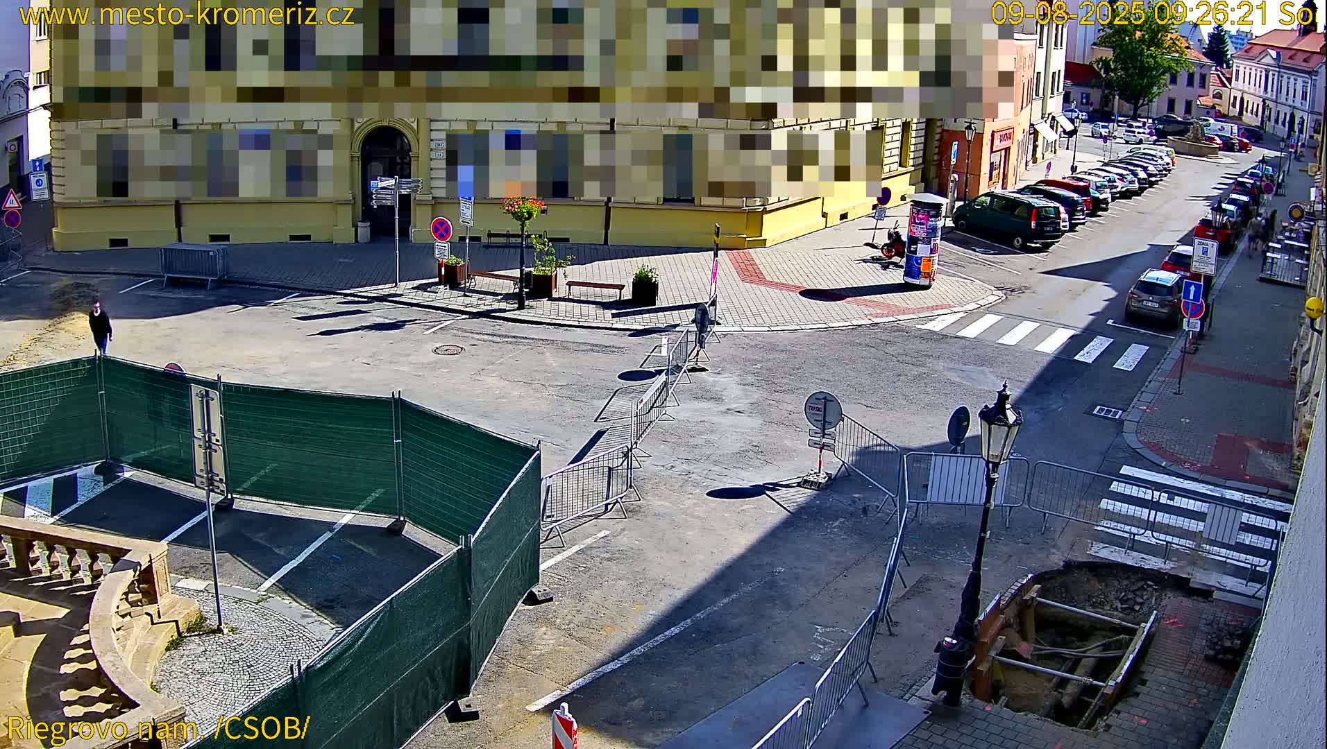 A sunny street scene shows a town square under construction, with temporary fencing, a person walking, and parked cars lining the street.