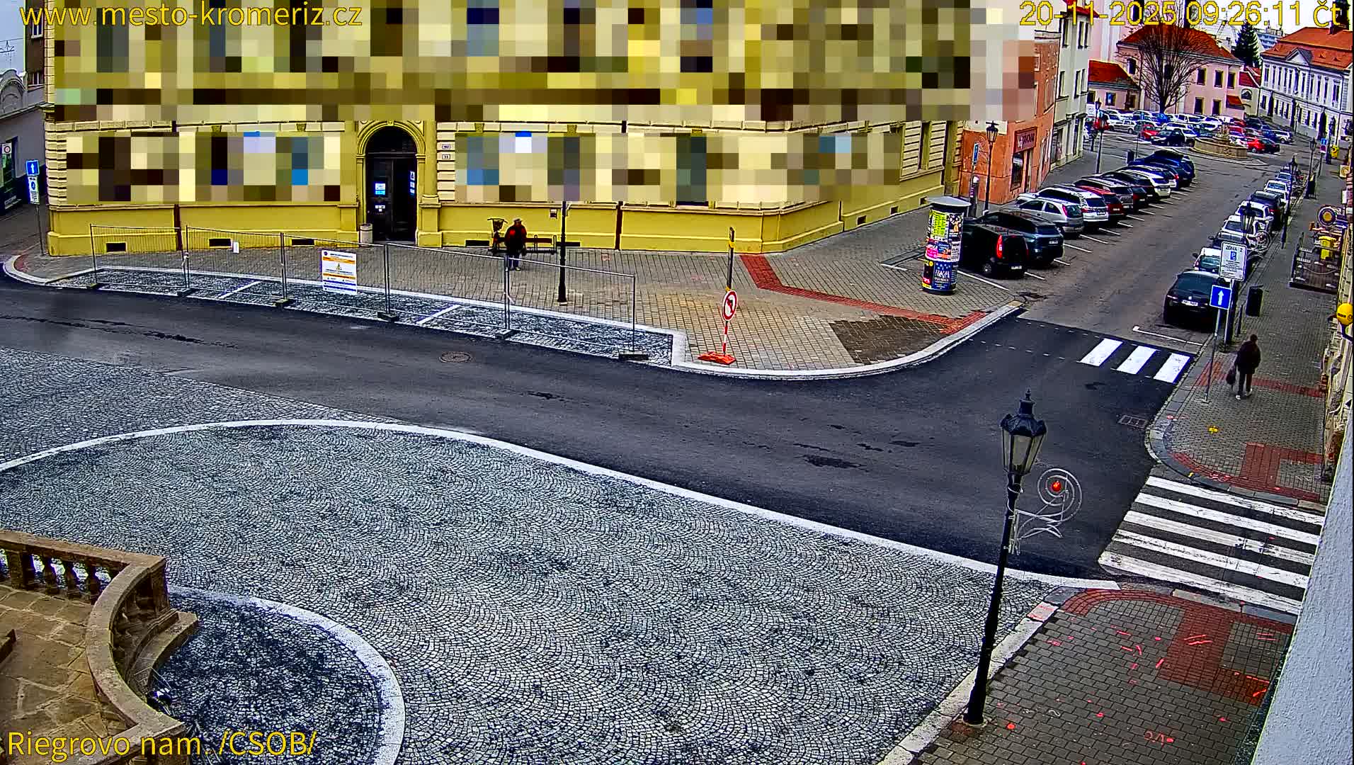 An elevated street-level view shows a quiet European town square featuring cobblestone paths, an asphalt road with a crosswalk, several parked cars, a prominent yellow building with pixelated windows, a construction barrier, and a few pedestrians, all under clear and dry conditions.