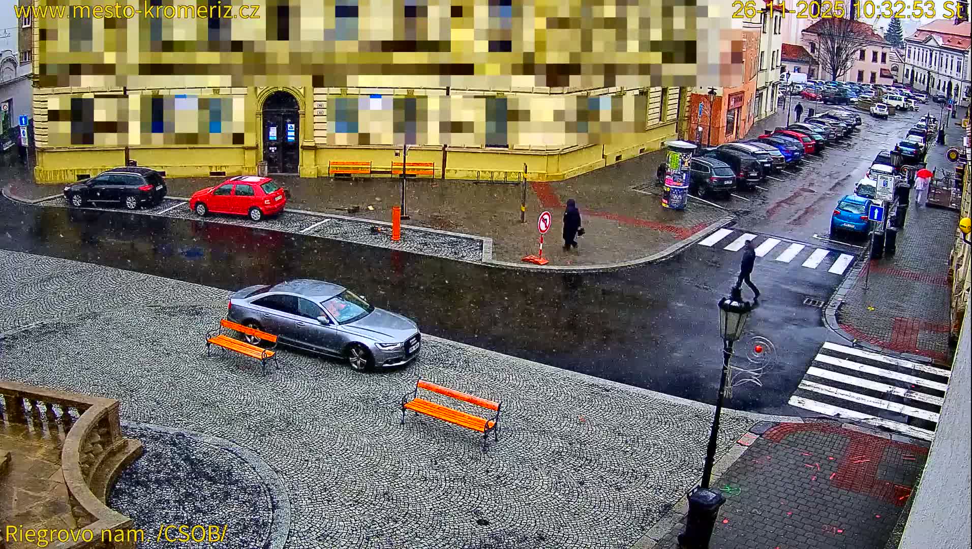 A high-angle view captures a rainy day on a cobblestone street in a European town, featuring numerous parked cars, several pedestrians, and historic buildings with wet, reflective surfaces.