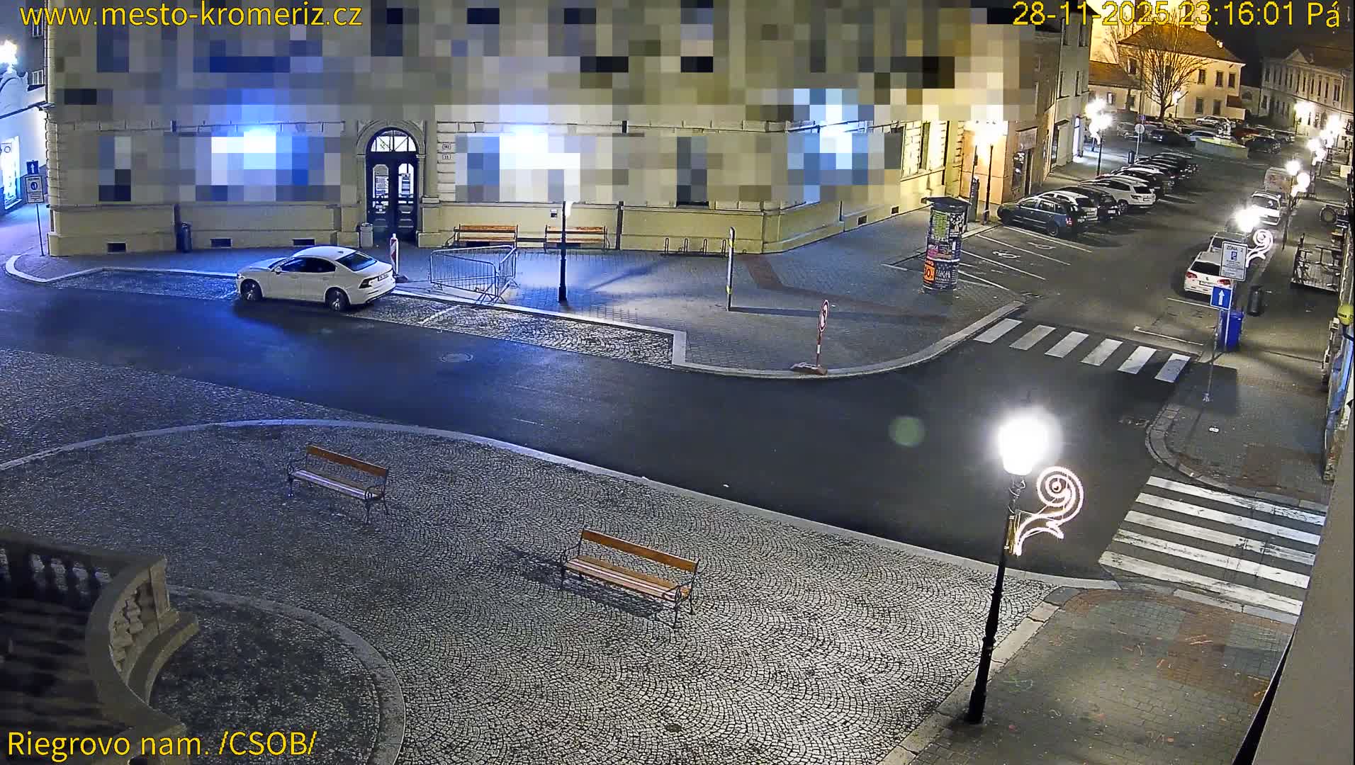 A nocturnal overhead view reveals a quiet European town square and street with a white car at a corner, parked vehicles, cobblestone sidewalks with benches, and illuminated streetlights under clear, dry night conditions.