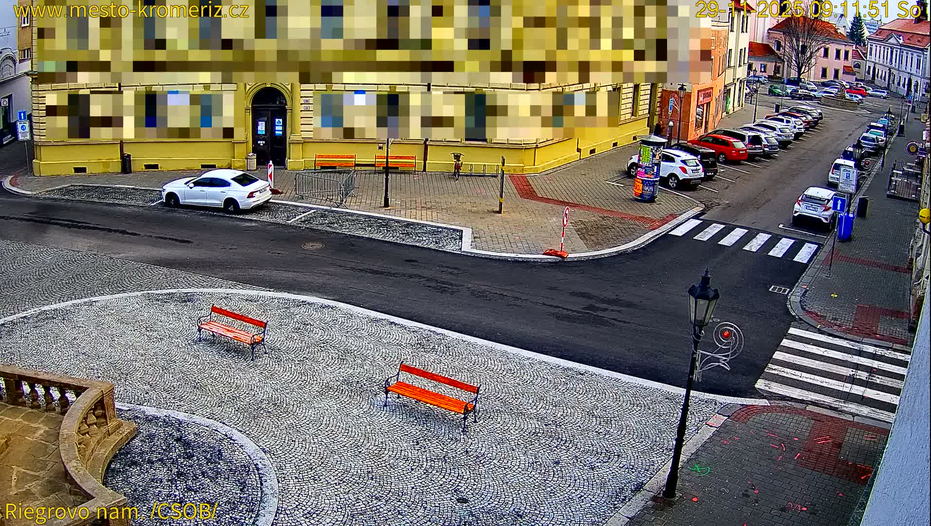 An elevated view reveals a quiet urban street and a cobblestone square with orange benches, lined by multistory buildings and various parked cars, under the even light of a mild day.