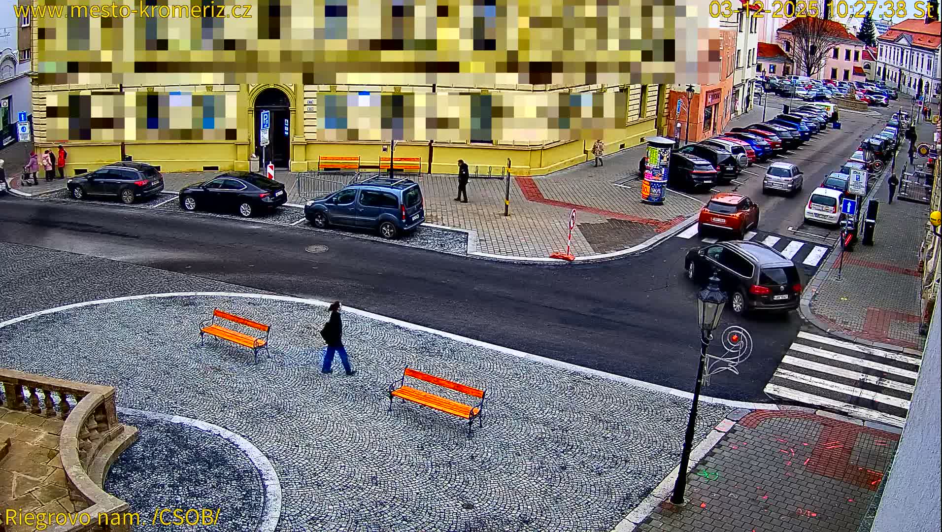An elevated wide shot reveals a European city street and square on an overcast day, featuring pedestrians walking on cobblestone paths, numerous parked cars, and a prominent yellow building.