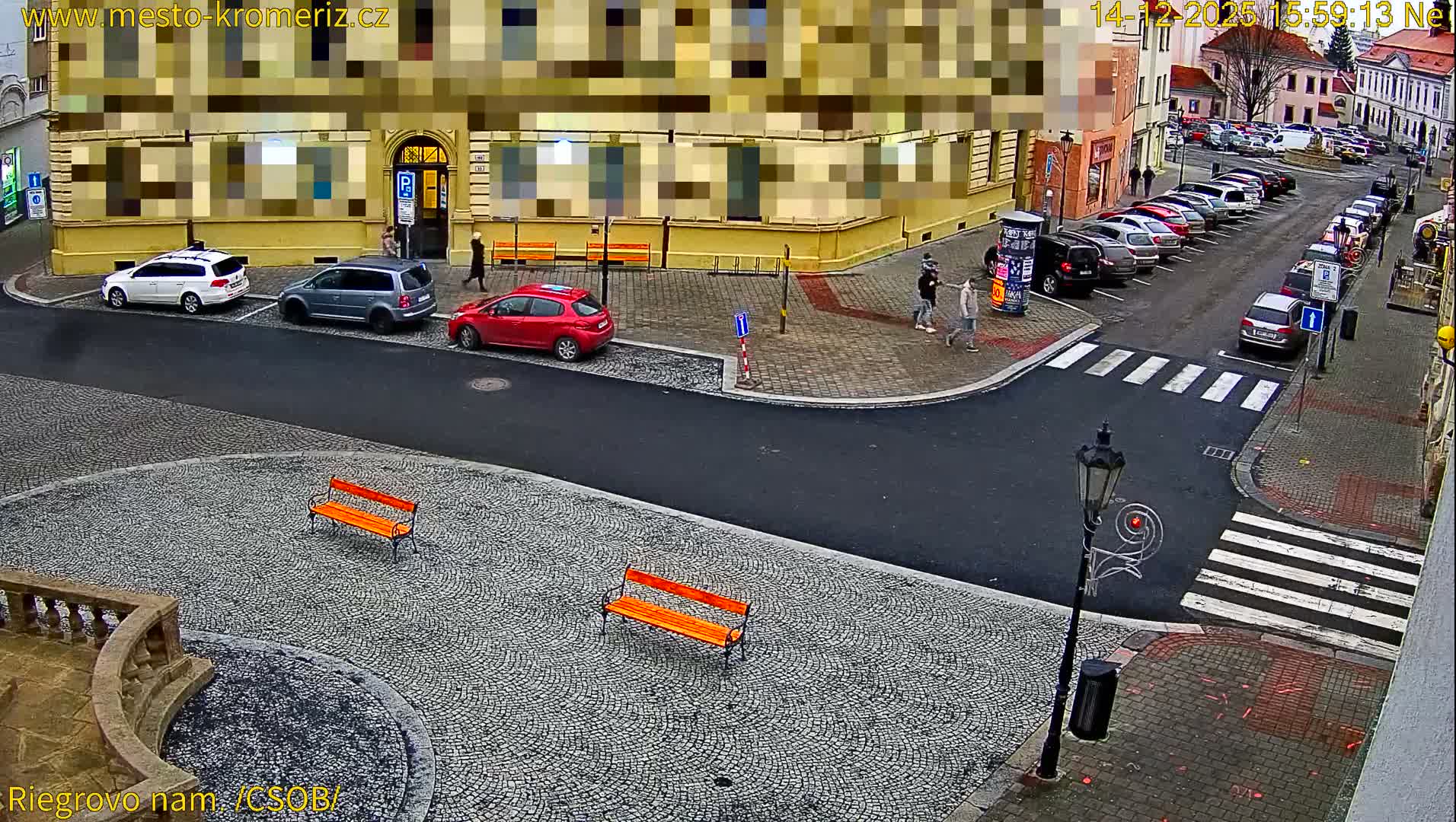 A damp urban street and square are seen at night, illuminated by bright streetlights reflecting off the wet pavement and roads, with several cars parked and buildings visible in the background.