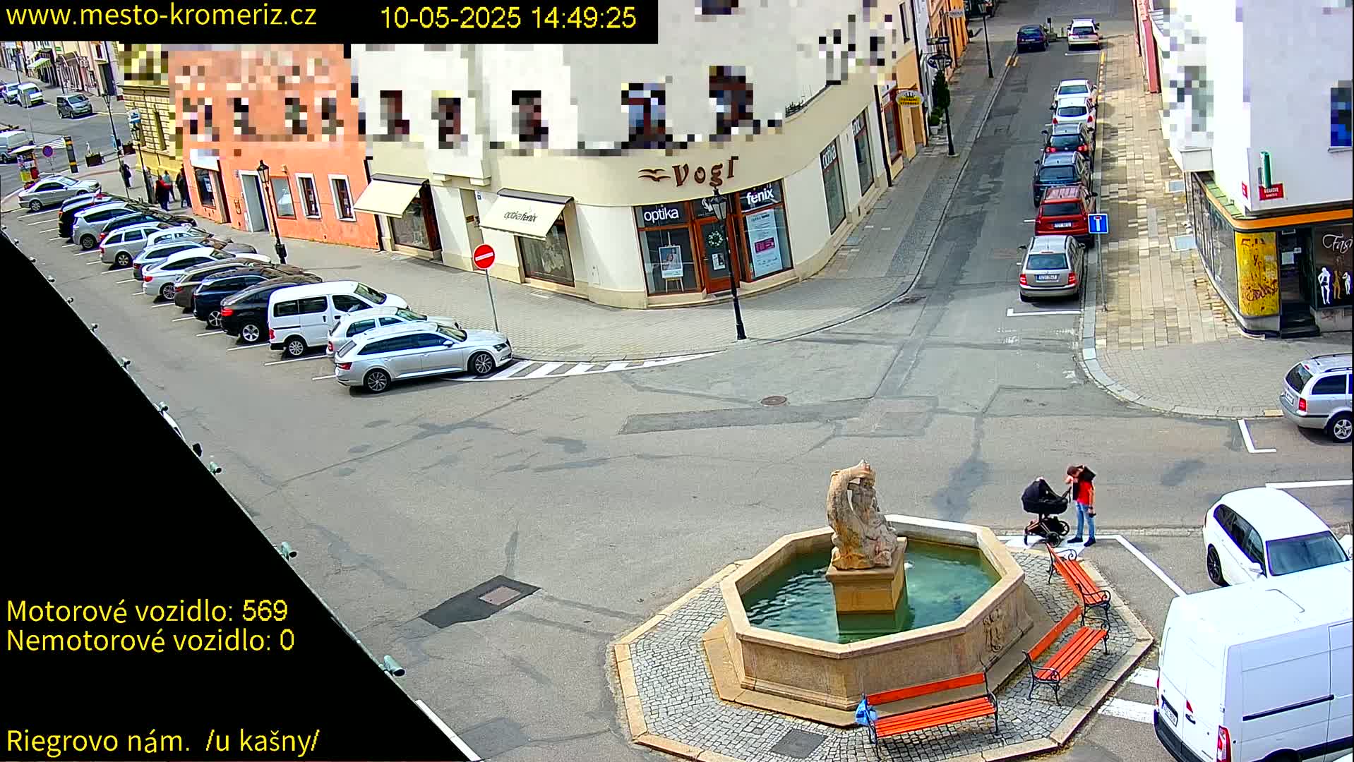 A town square with a stone fountain, several parked cars, and two people with a stroller, on a sunny day.