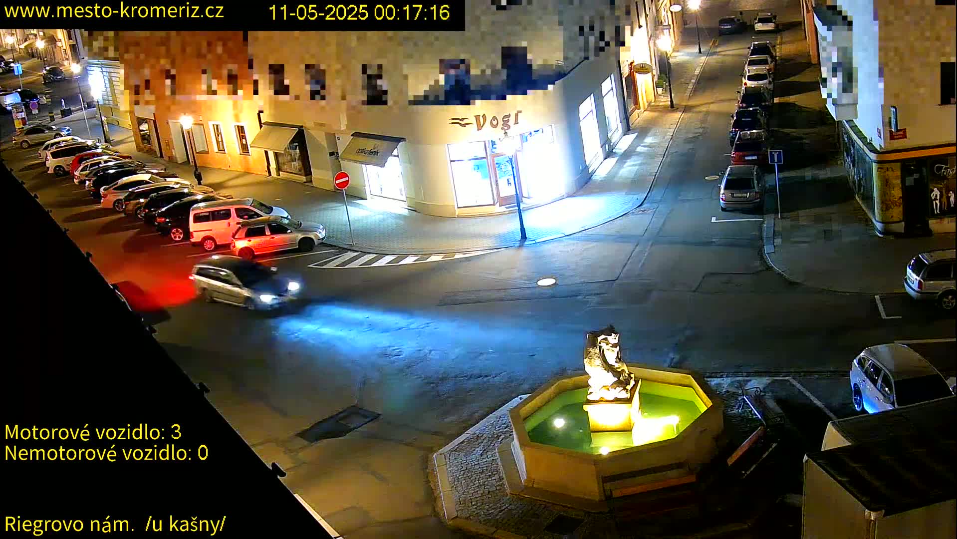 At night, several cars are parked along the side of a street next to buildings, with a fountain in the center of the intersection and a few cars driving by.