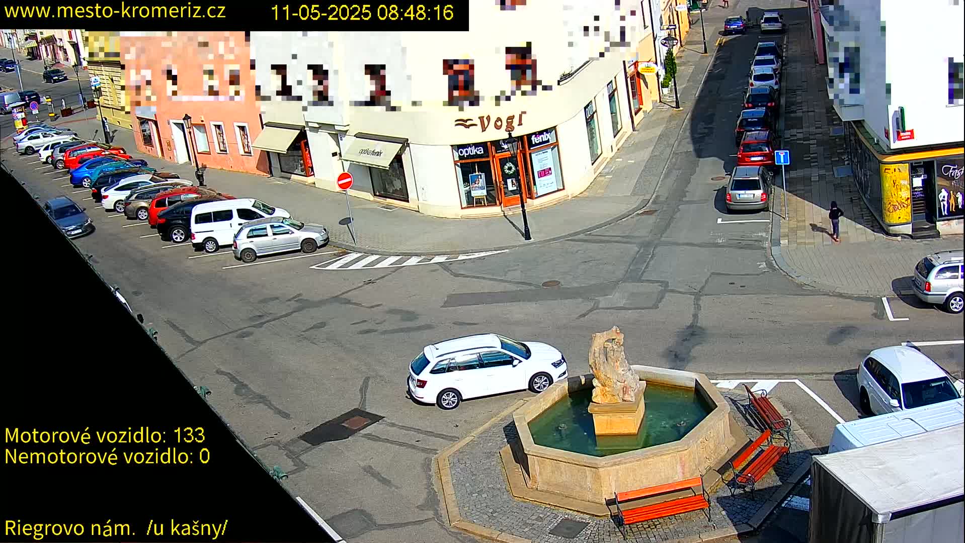 A sunny town square scene shows several parked cars lining a street, a white station wagon driving away from a stone fountain with benches around it.