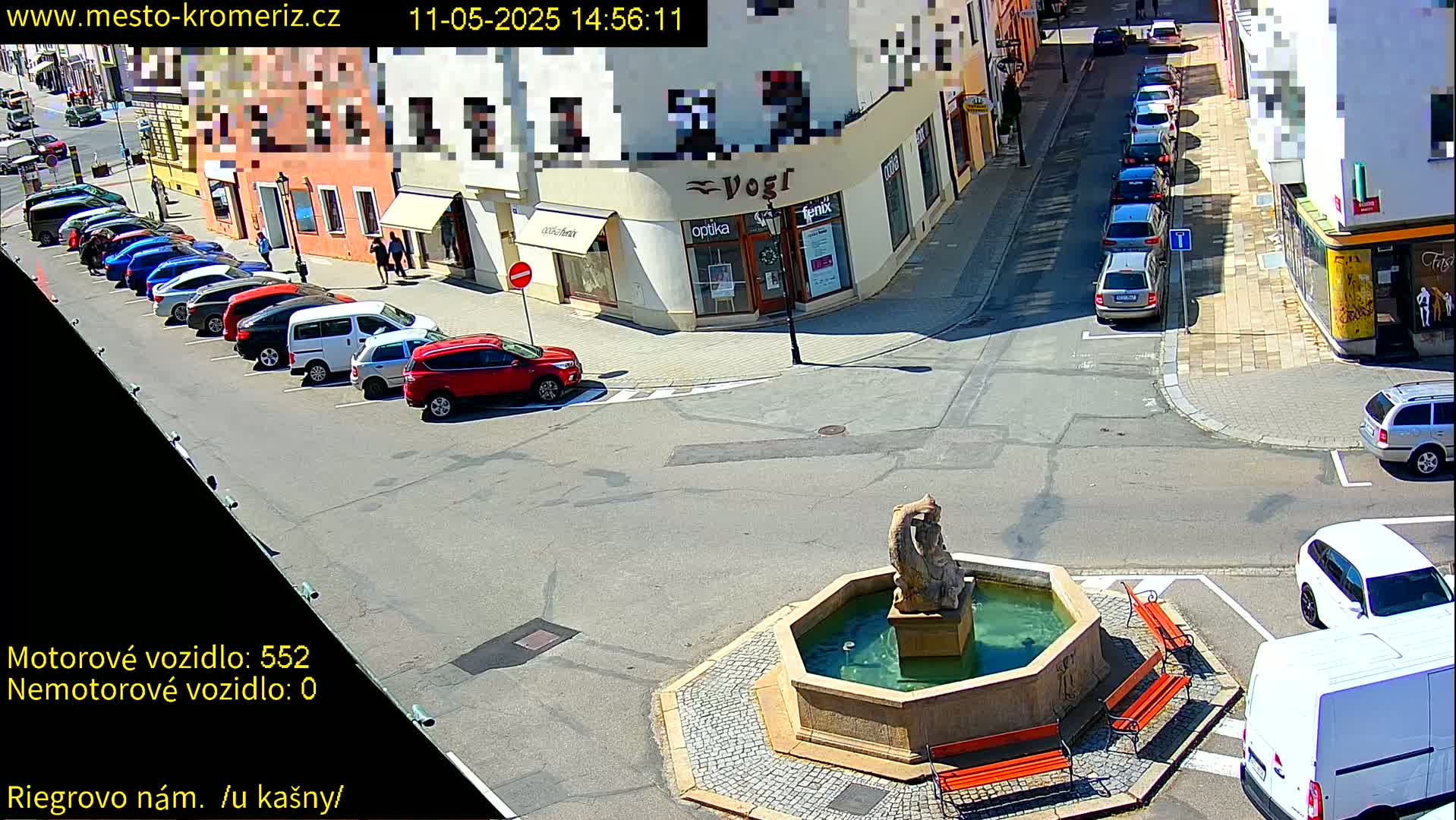 A sunny street scene shows several cars parked along the side of a street and others driving by a stone fountain in a town square with several benches surrounding it.