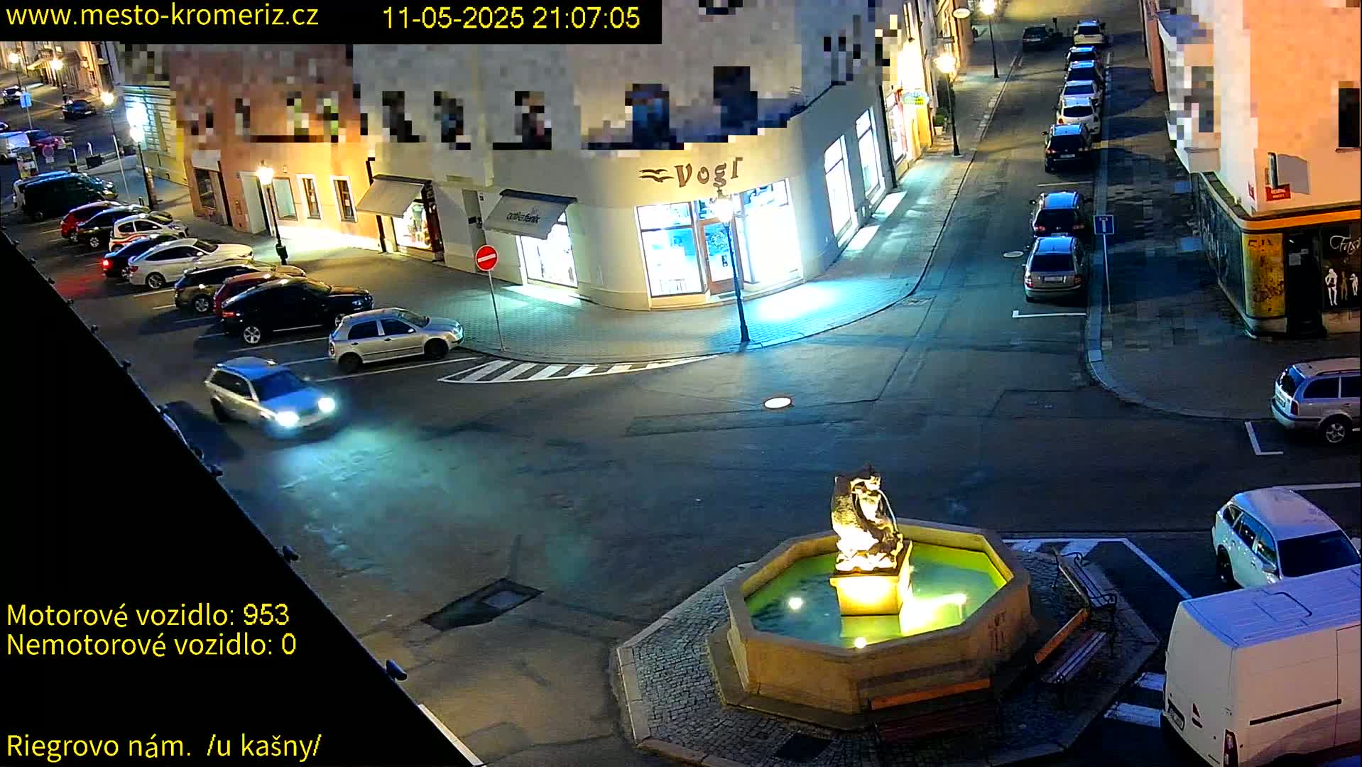A nighttime, street-level view of a town square with several parked cars surrounding a fountain featuring a statue.