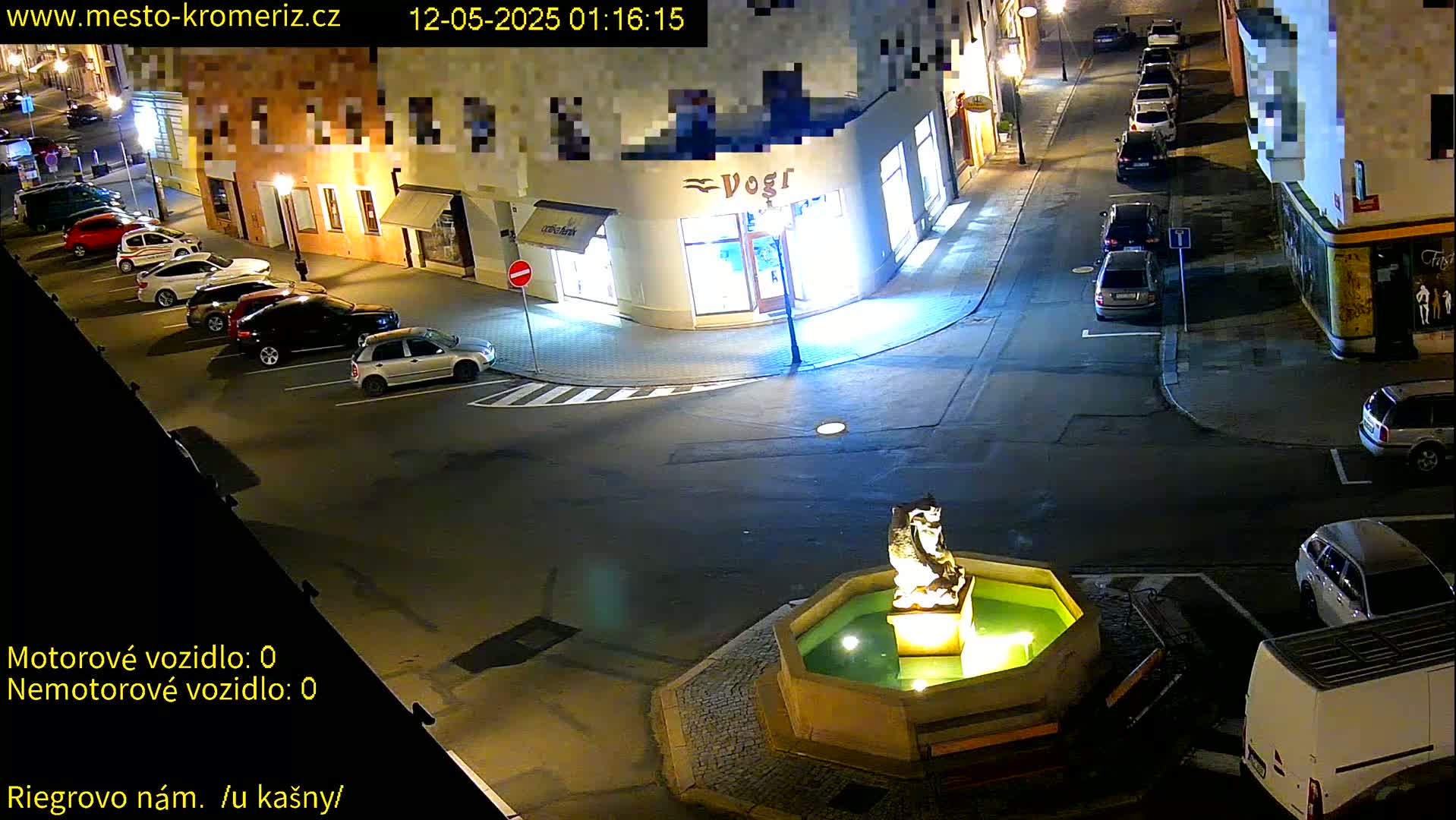 A nighttime aerial view of a town square shows several parked cars surrounding a fountain with a statue, under partly lit streetlights.