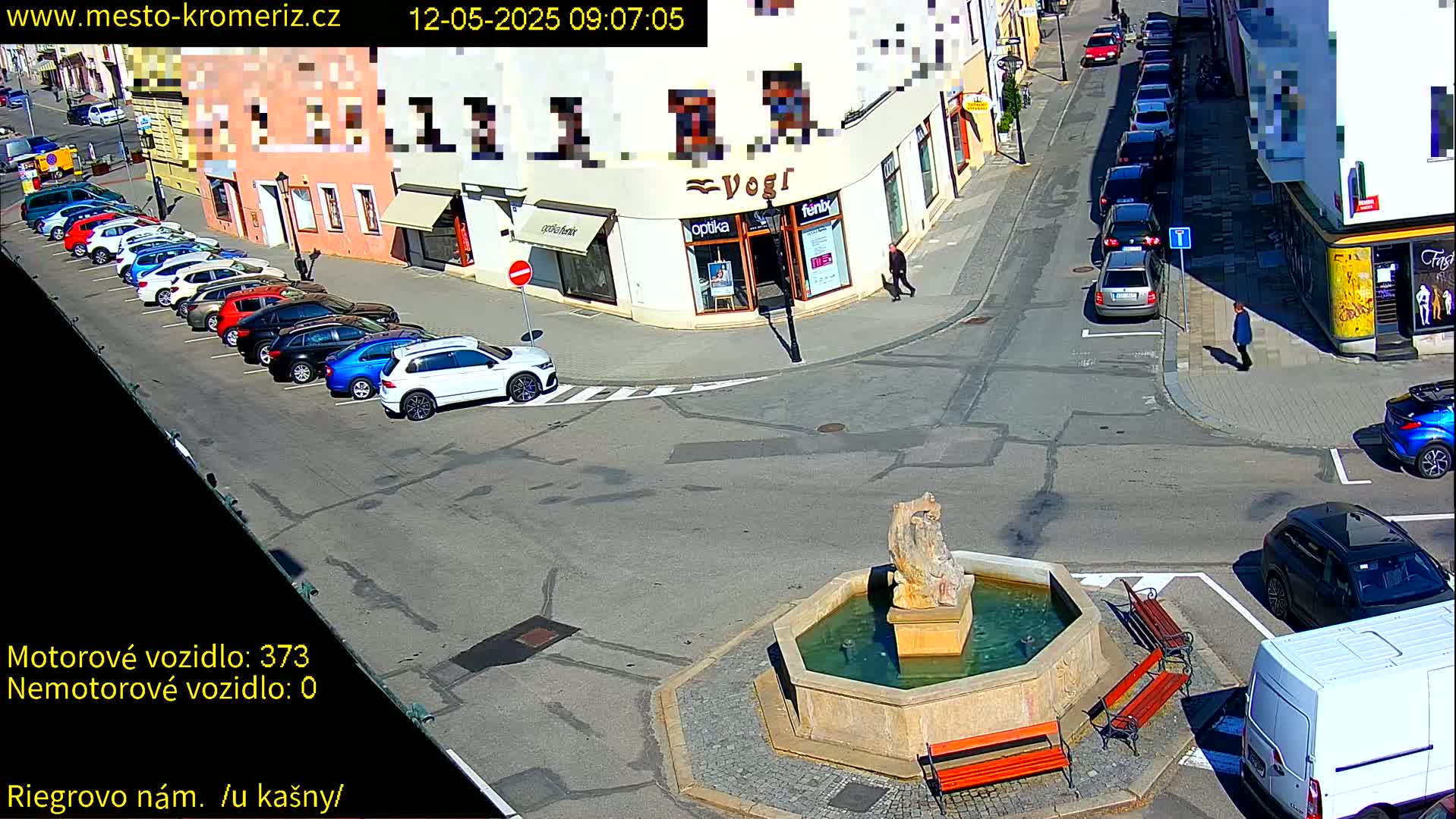 A sunny street scene shows a fountain in a town square with parked cars lining the sides of the street, and a few pedestrians walking by.