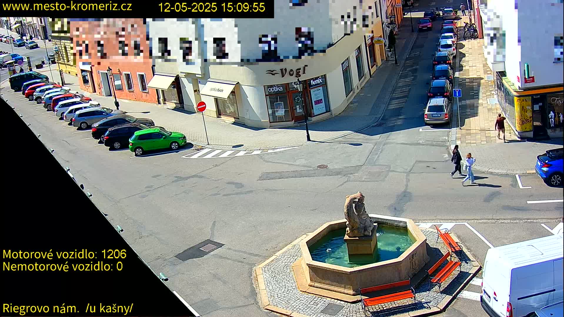 A sunny town square features a fountain with benches, several parked cars, and pedestrians crossing a street.