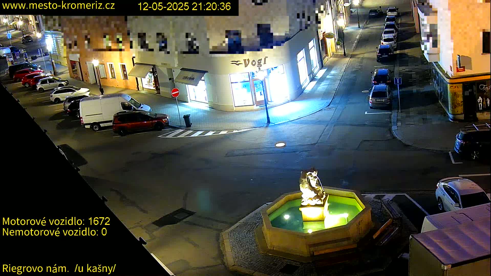 A nighttime street scene shows a fountain with a statue in the center of a plaza, surrounded by parked cars and buildings.