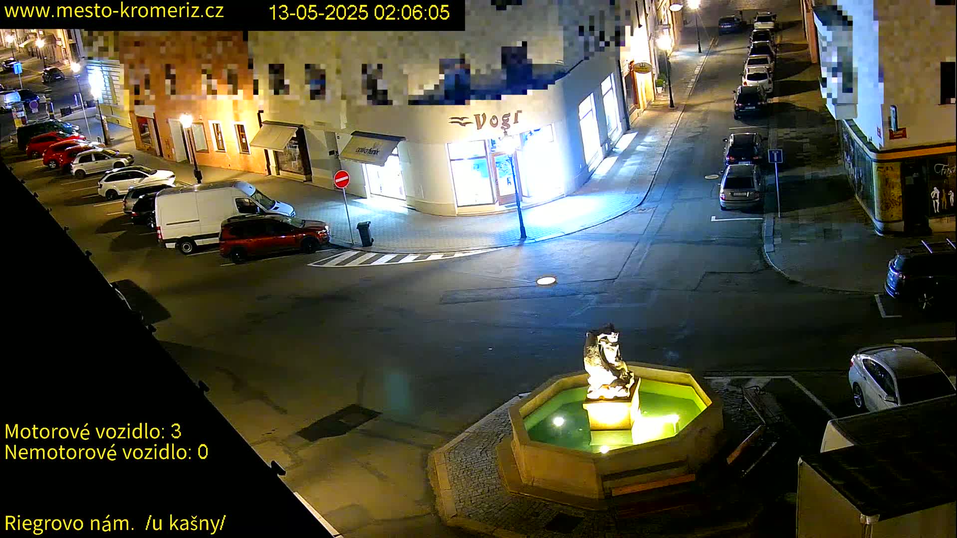 A nighttime street scene shows several parked cars surrounding a stone fountain with a statue in a town square.