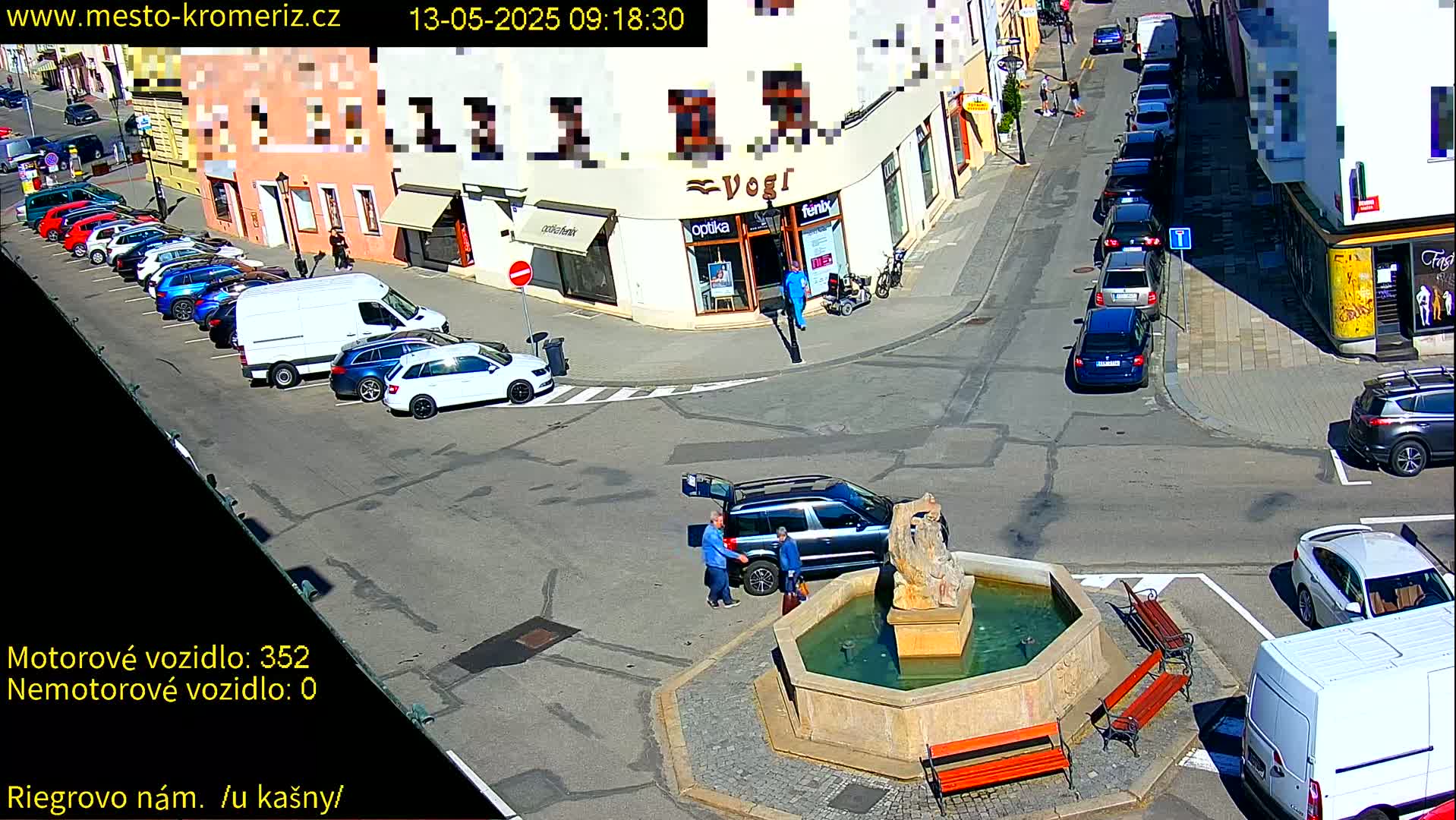 A sunny town square with a fountain, several parked cars, and two people standing near a vehicle with its trunk open.