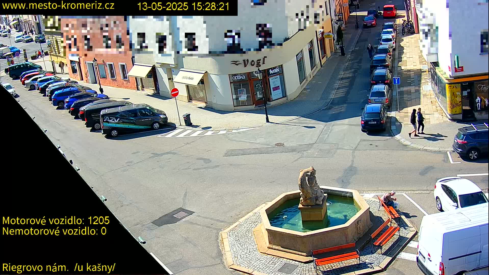 A sunny town square features a central fountain, several parked cars, and pedestrians walking near shops.