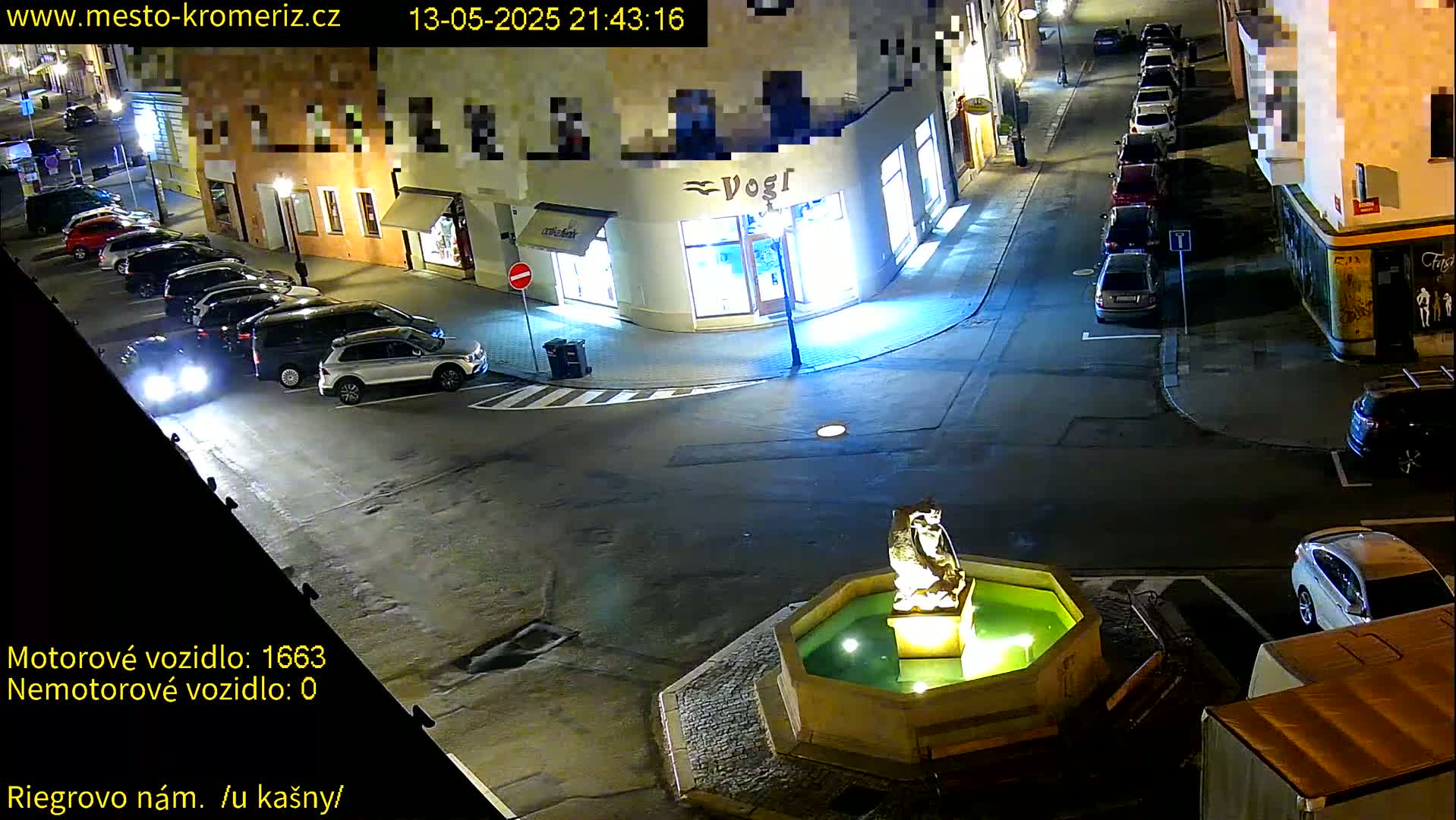 A nighttime street scene shows several parked cars surrounding a fountain with a statue in the center, under clear skies.