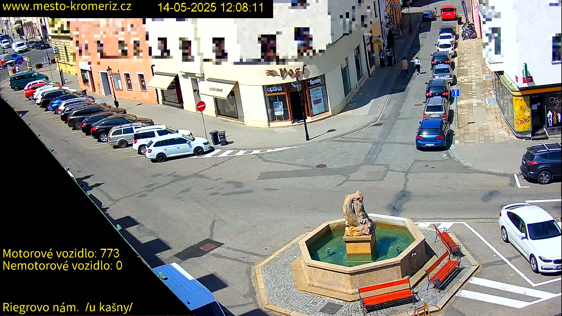 A sunny town square features a stone fountain, several parked cars, and buildings with awnings.