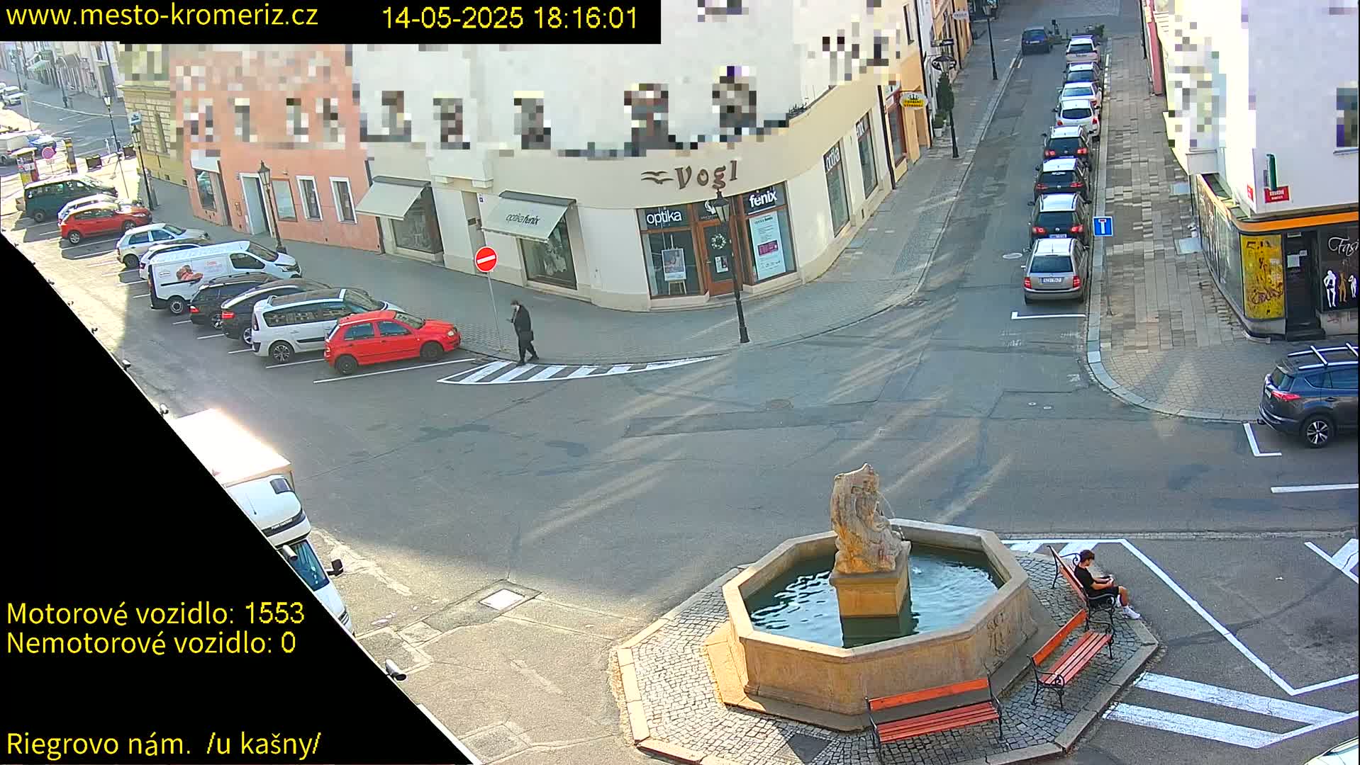 A sunny street scene shows several parked cars, a pedestrian, and a person sitting on a bench by a stone fountain in a town square.