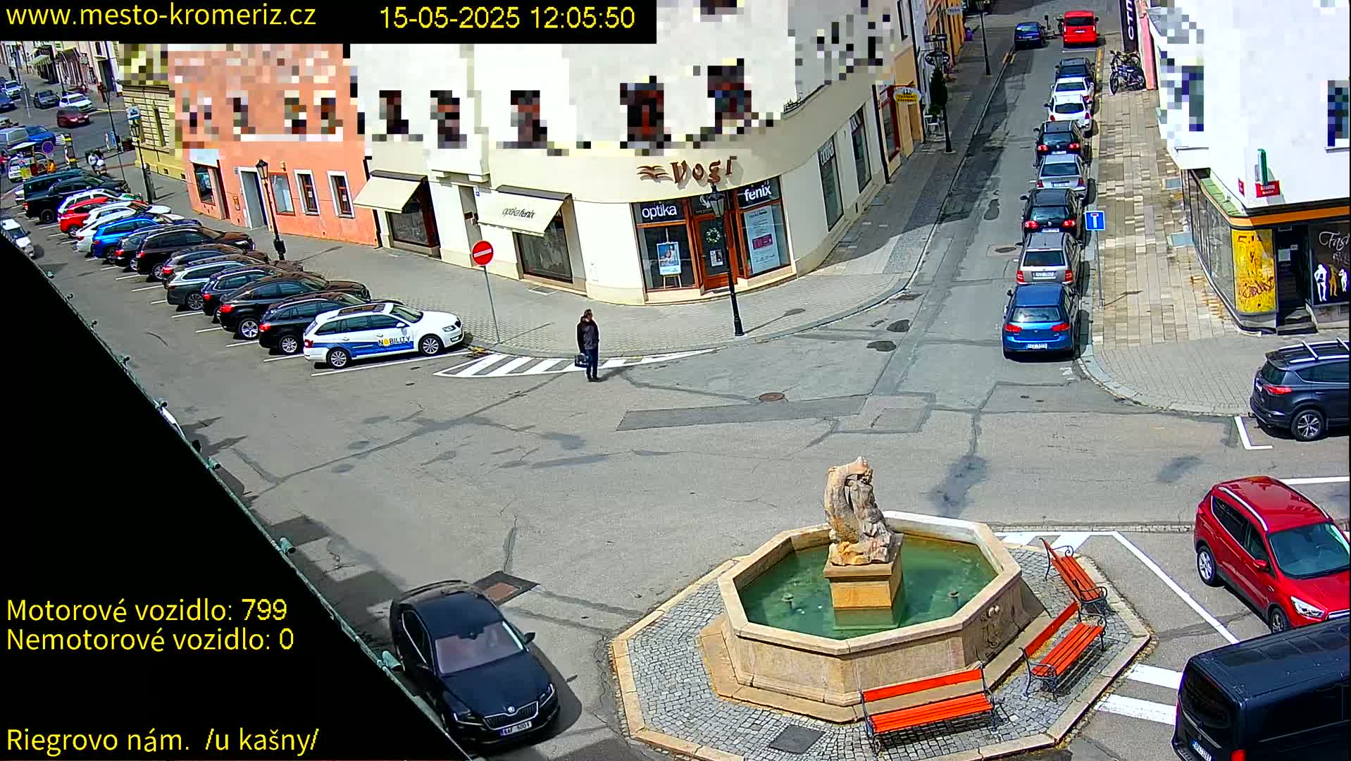 A sunny town square scene shows a fountain, several parked cars, a police car, and a pedestrian, with additional cars driving on adjacent streets.