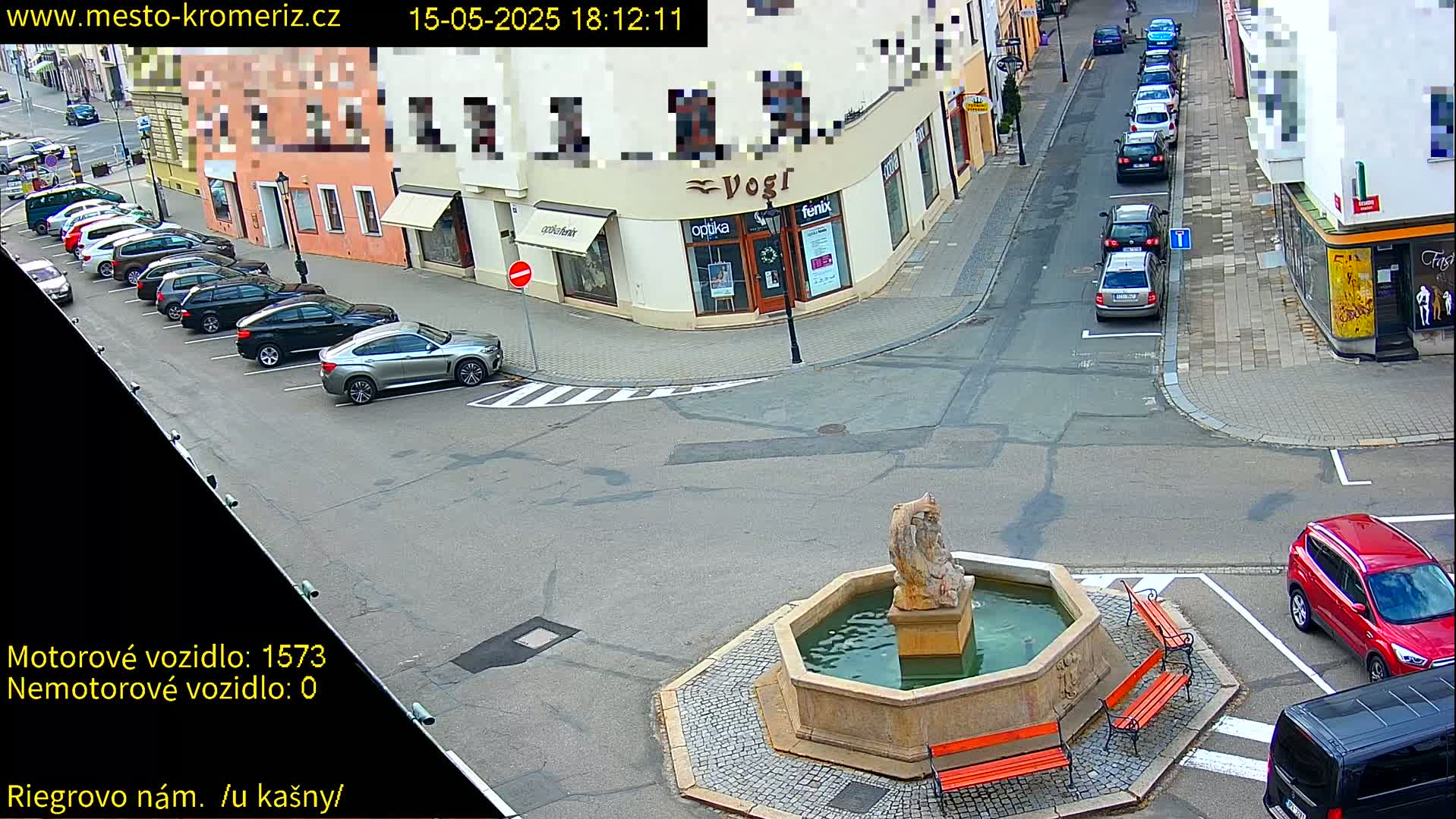 A stone fountain with a statue sits in a town square surrounded by parked cars and buildings, under a clear sky.