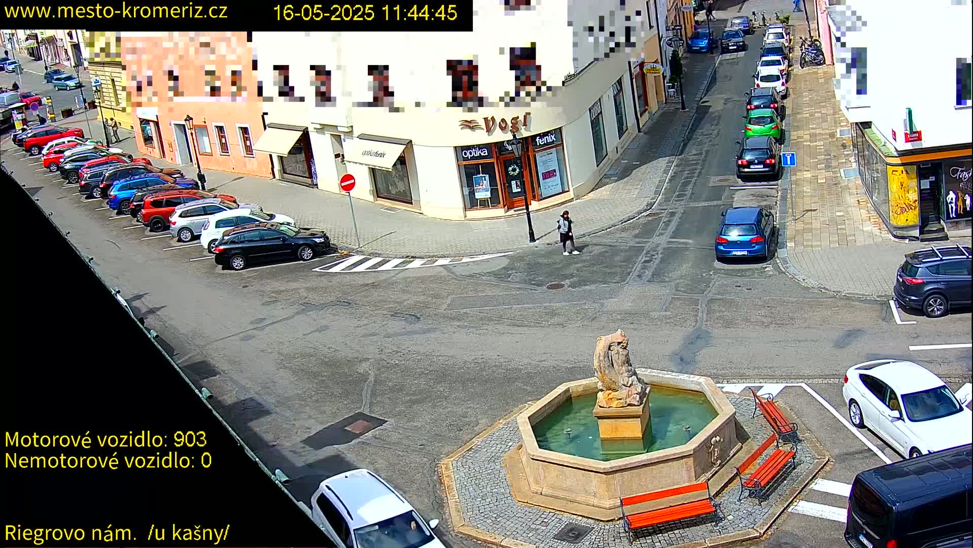 A sunny town square features a fountain with several benches surrounding it, cars parked along the street, and a pedestrian walking near buildings.