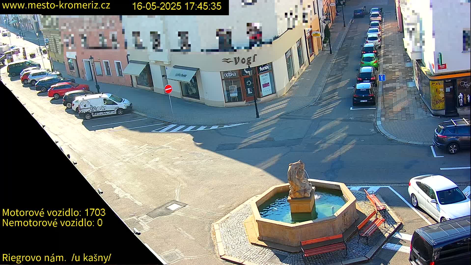A sunny town square features a stone fountain with several benches surrounding it, and cars parked along the surrounding streets.