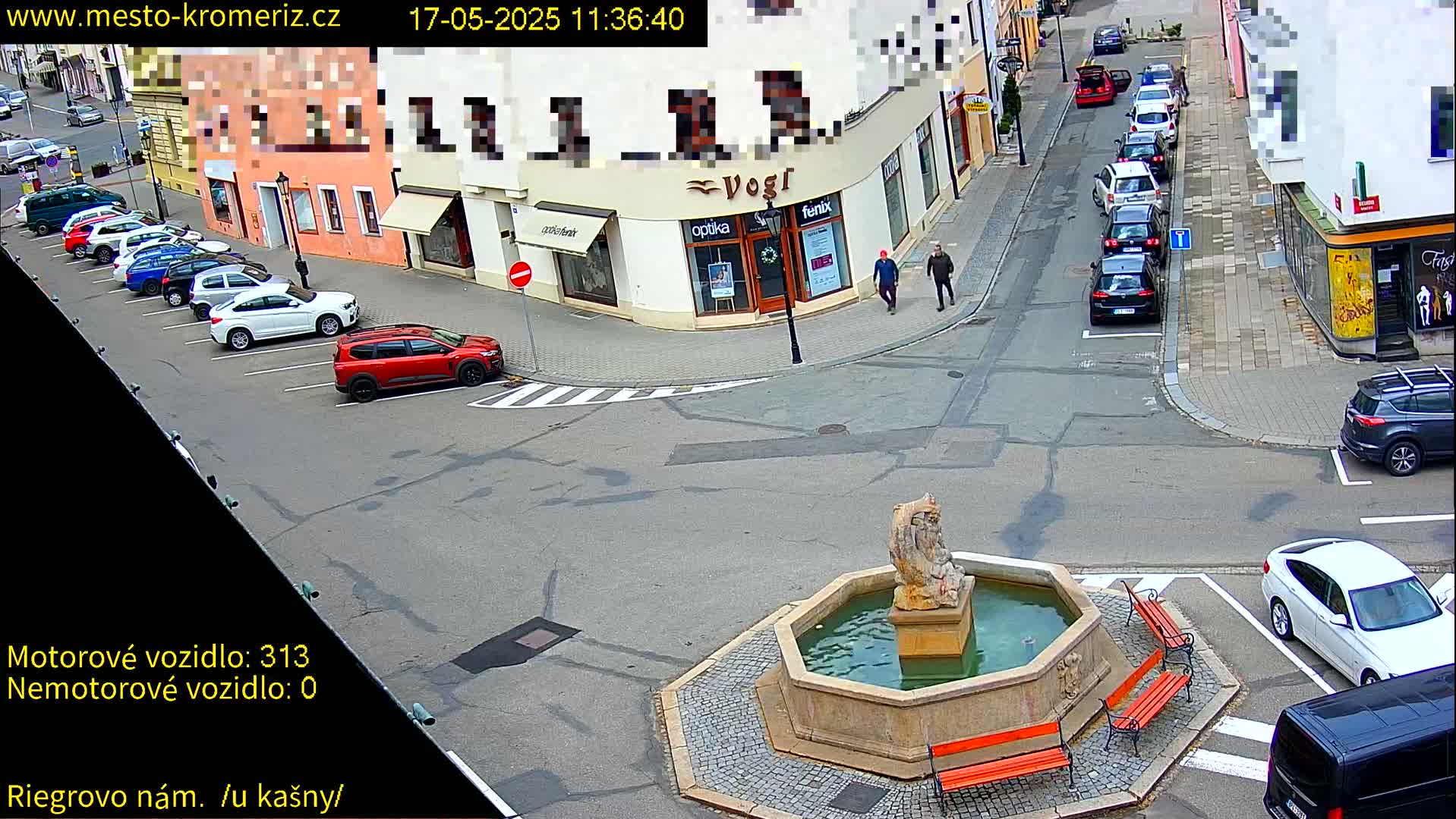 A street scene shows a town square with a stone fountain, several parked cars, and a few pedestrians walking near shops on a sunny day.