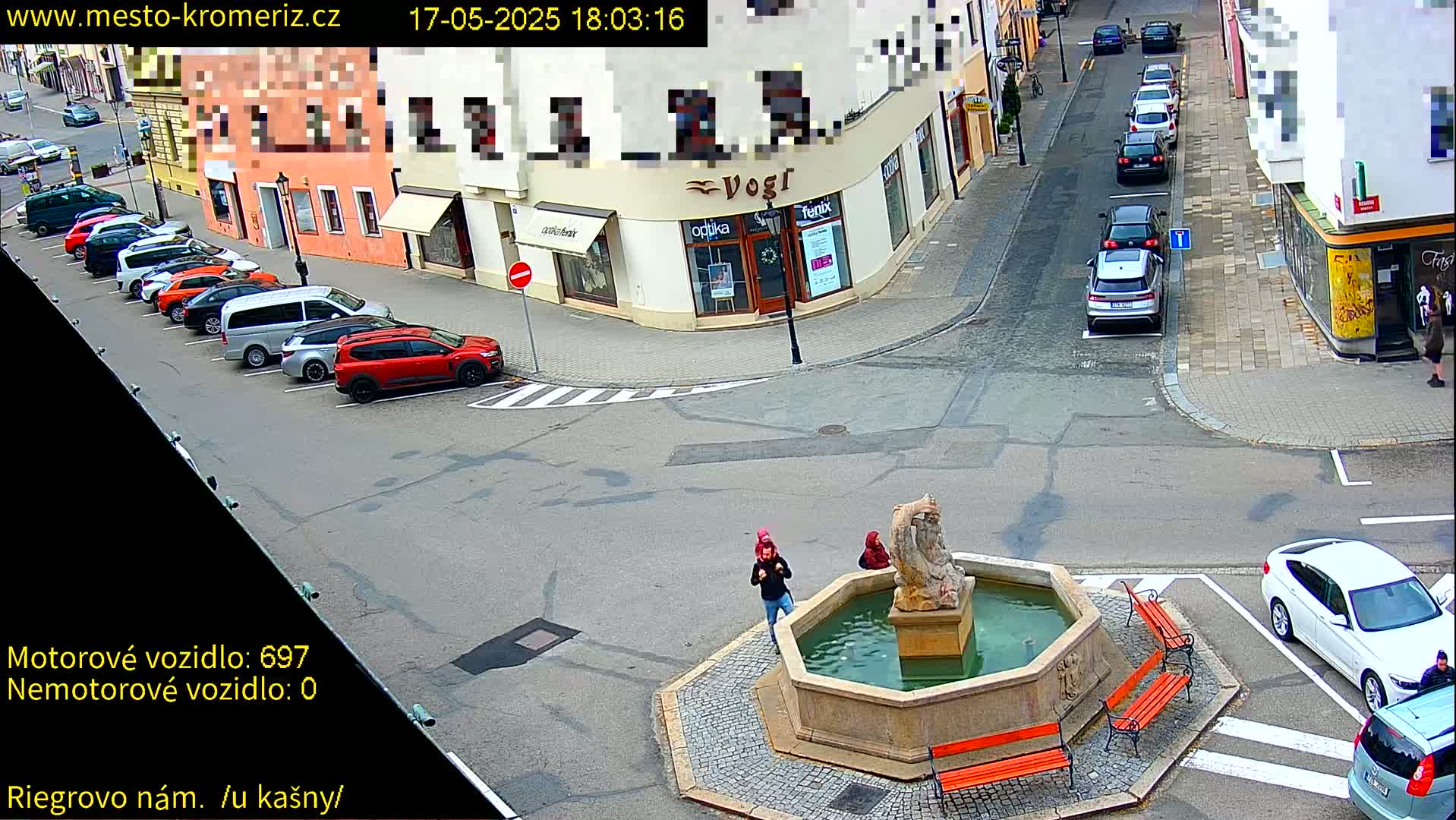 A town square with a fountain, several parked cars, and a few pedestrians, on a sunny day.