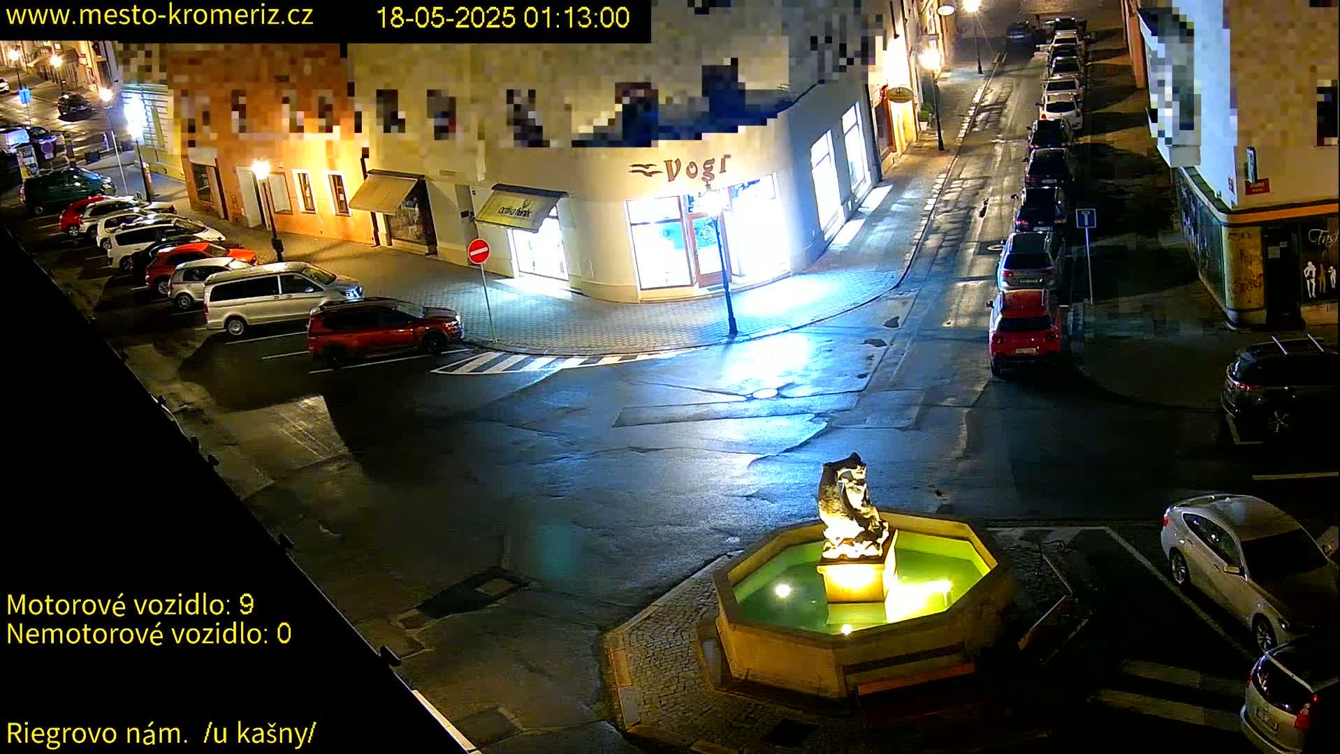 A nighttime street scene shows several parked cars around a fountain with a statue, illuminated by streetlights.