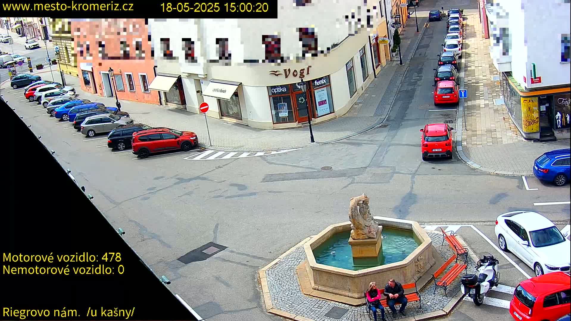 A sunny town square features a stone fountain, several parked cars, and two people sitting on a bench.