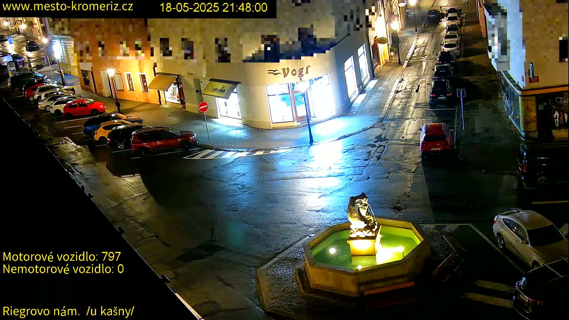 A wet, nighttime street scene shows several cars parked along the sides of buildings surrounding a small fountain with a statue in the center.