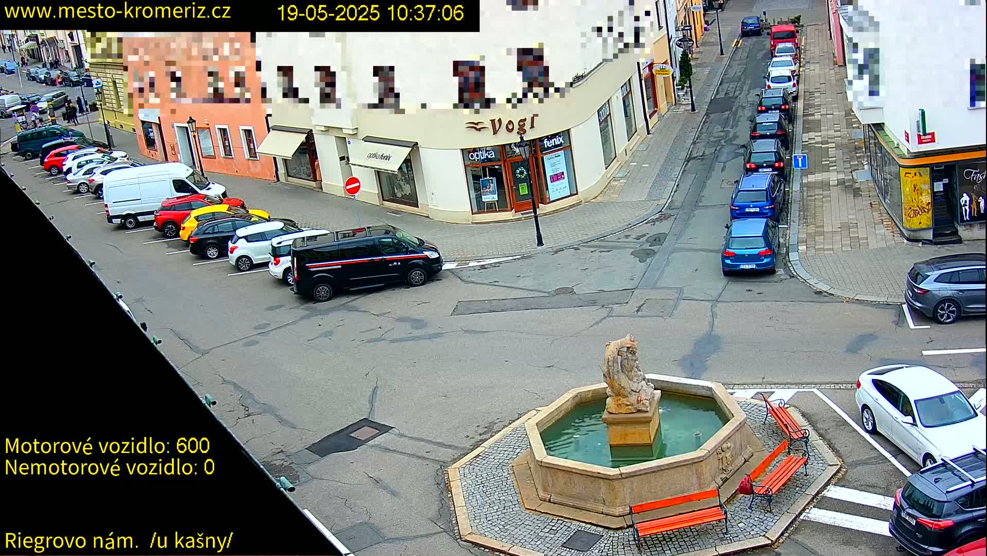 A town square with a stone fountain, surrounded by parked cars and buildings on a sunny day.