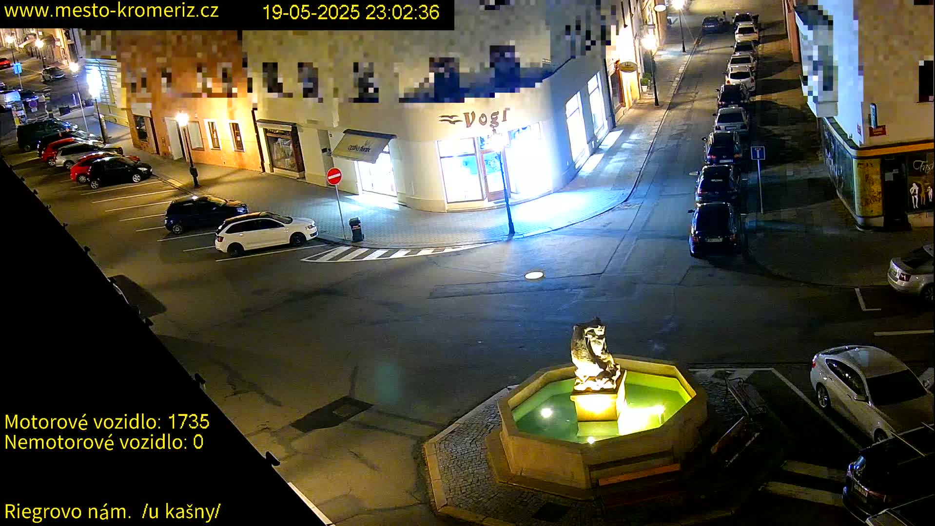 A nighttime, street-level view of a town square with parked cars, a fountain featuring a stone statue, and several buildings illuminated under artificial light.