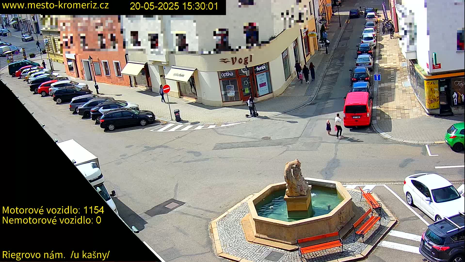 A sunny town square with a fountain at its center, surrounded by parked cars, pedestrians, and buildings.