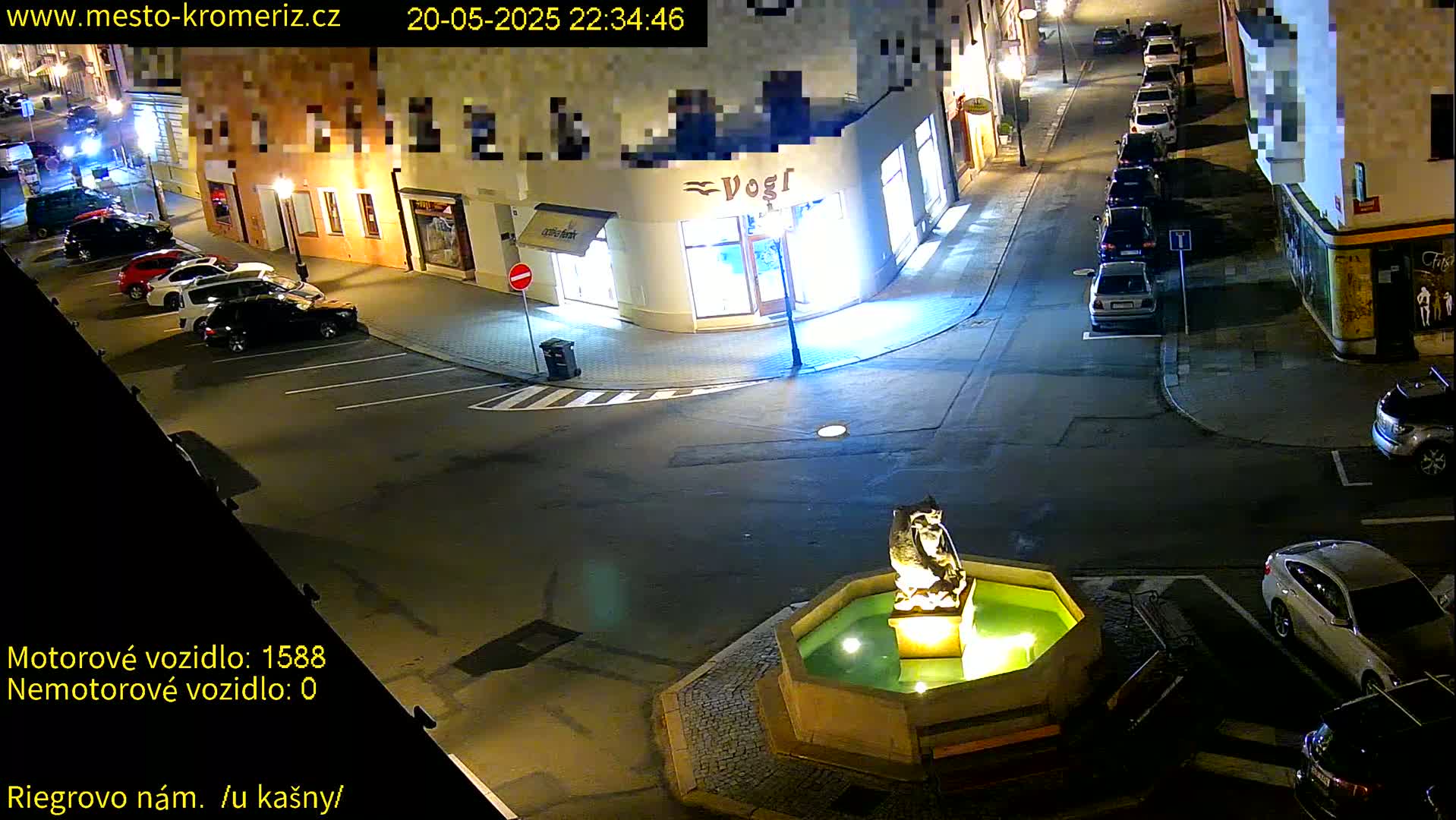 A nighttime street scene shows several parked cars surrounding a stone fountain in a town square.
